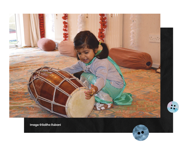  A very young girl in a colourful kneels down to play with large drum instrument and a young woman makes notes intently in front of images hung in a gallery 