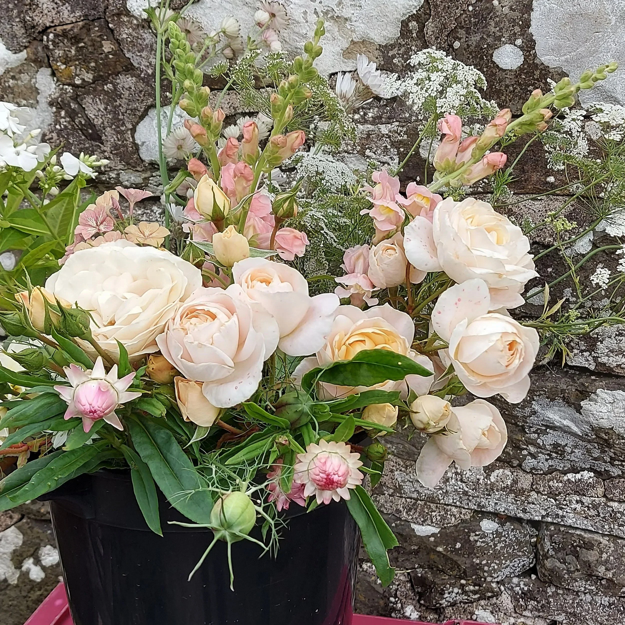 A black pot filled with light pink and white roses and other assorted flowers and greenery, placed in front of a stone wall.