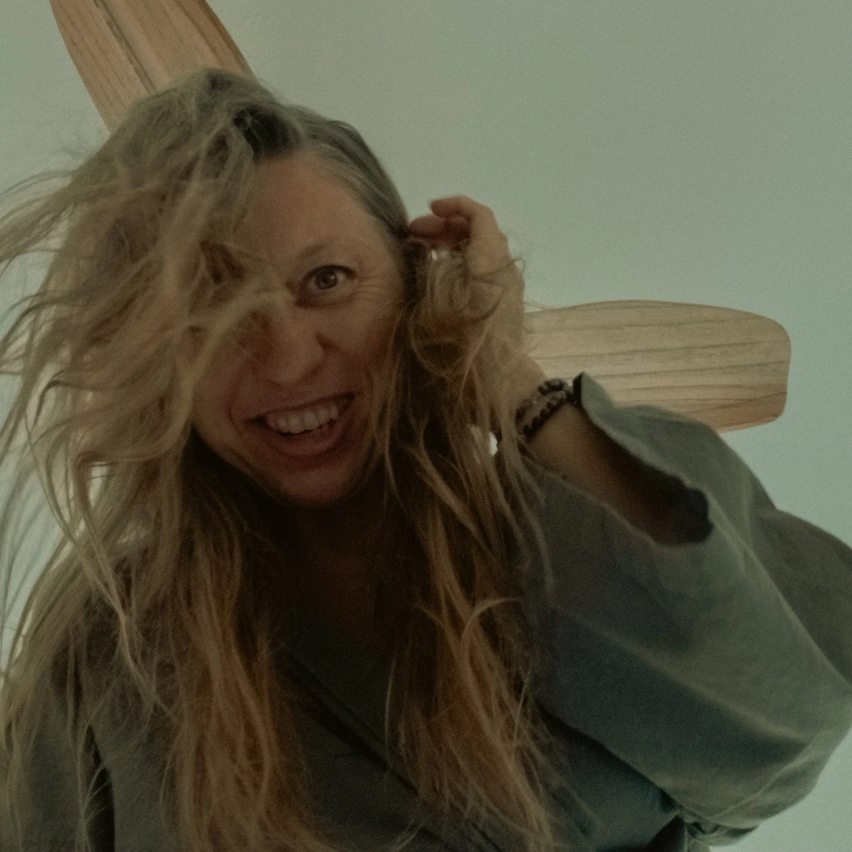 A woman with long, curly blond hair smiling and holding her hair with her left hand, wearing a casual jacket and a bracelet.