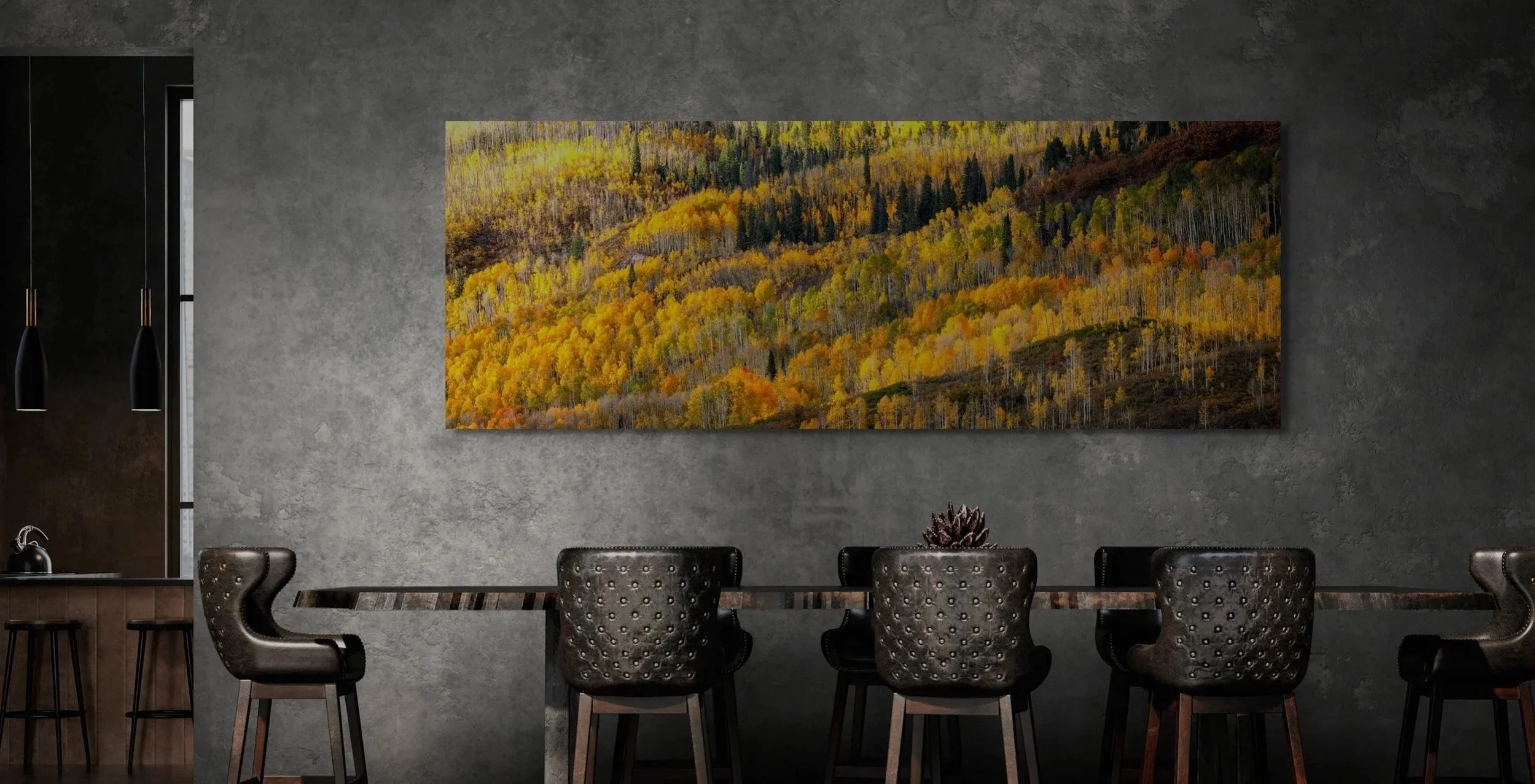 Interior of a modern dining area with a long table, black suede chairs, a dark wood countertop on the left, a black table lamp, and a large landscape painting of a colorful autumn forest wall.