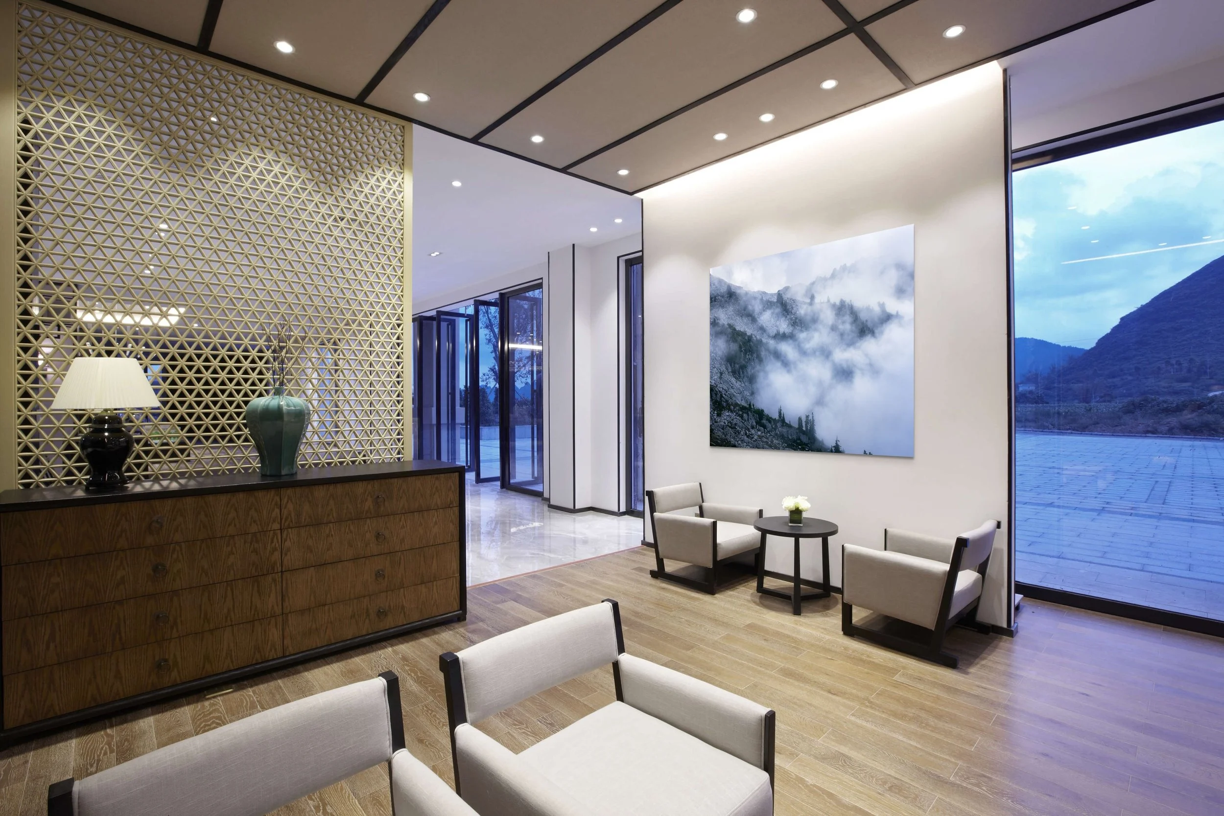 Modern lobby with wood flooring, white walls, and large windows showing mountains and water outside. There are chairs around small tables, a wooden sideboard with a lamp and a vase, and a large landscape photograph on the wall.