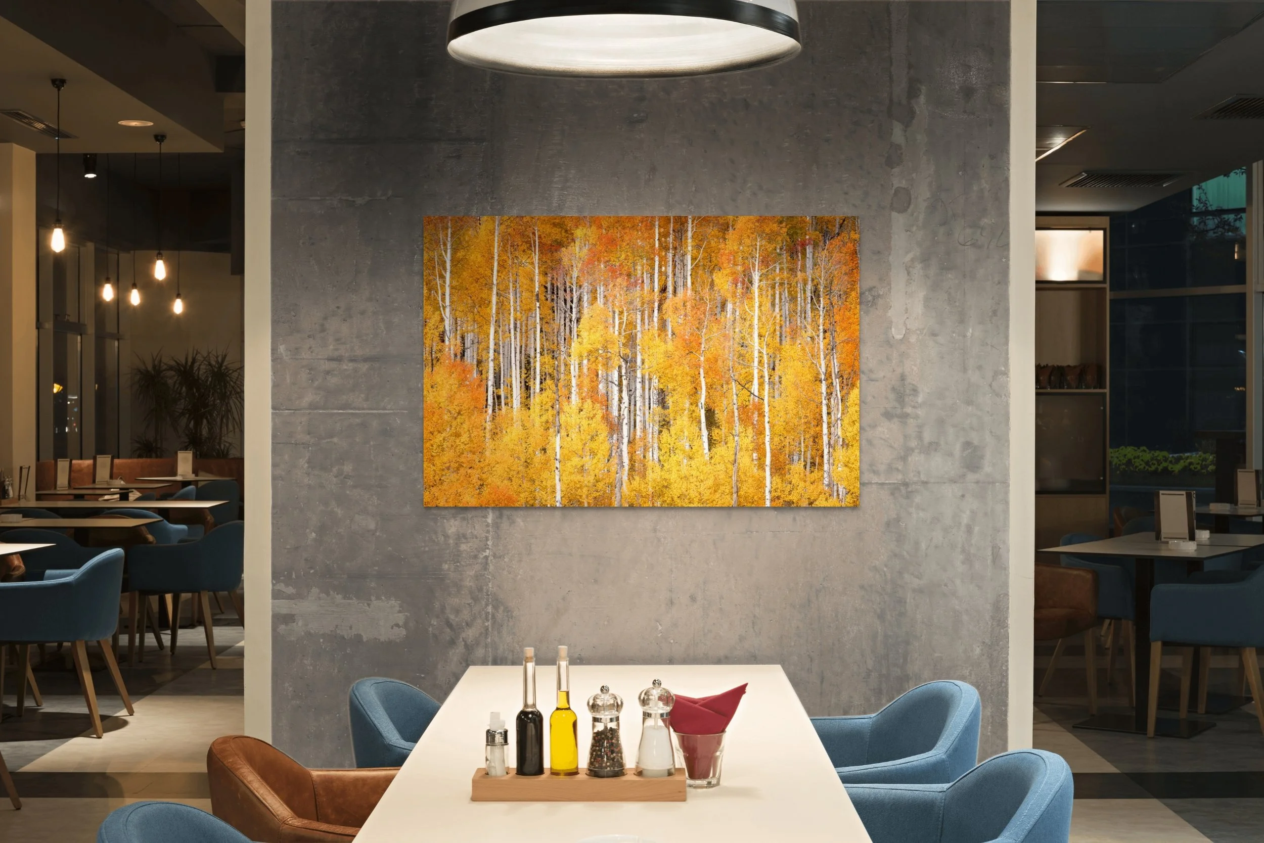 Interior of a modern restaurant with a concrete wall, colorful autumn landscape painting, white table with condiments, and blue and brown chairs.