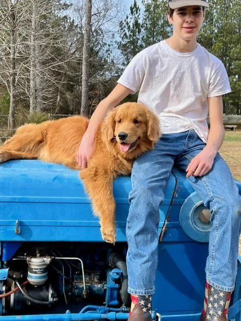 Young person wearing a white shirt and jeans sitting on a blue tractor with a Golden Retriever dog lounging next to them, outdoors in a rural setting.