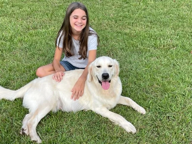 A girl sitting on grass next to a large white dog, both appear happy and relaxed.