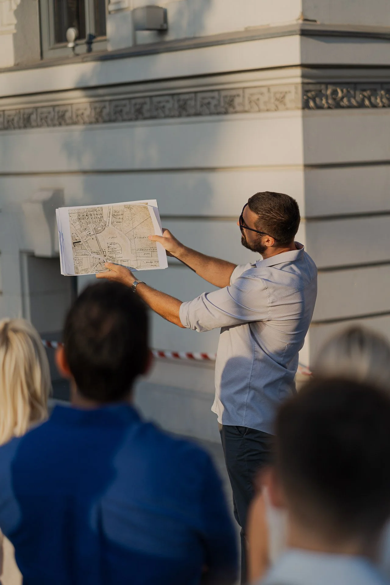 Man giving a presentation outdoors holding architectural plans, audience watching.