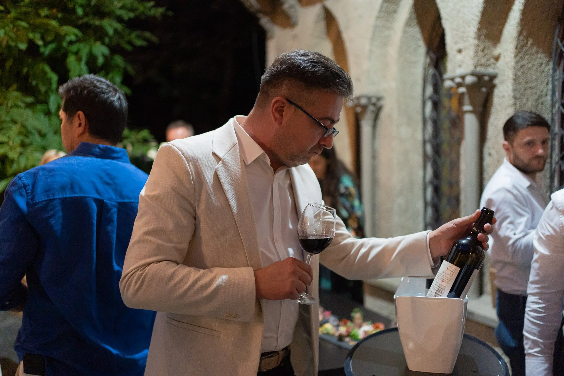 Man in a beige suit with glasses pouring red wine at a social gathering.