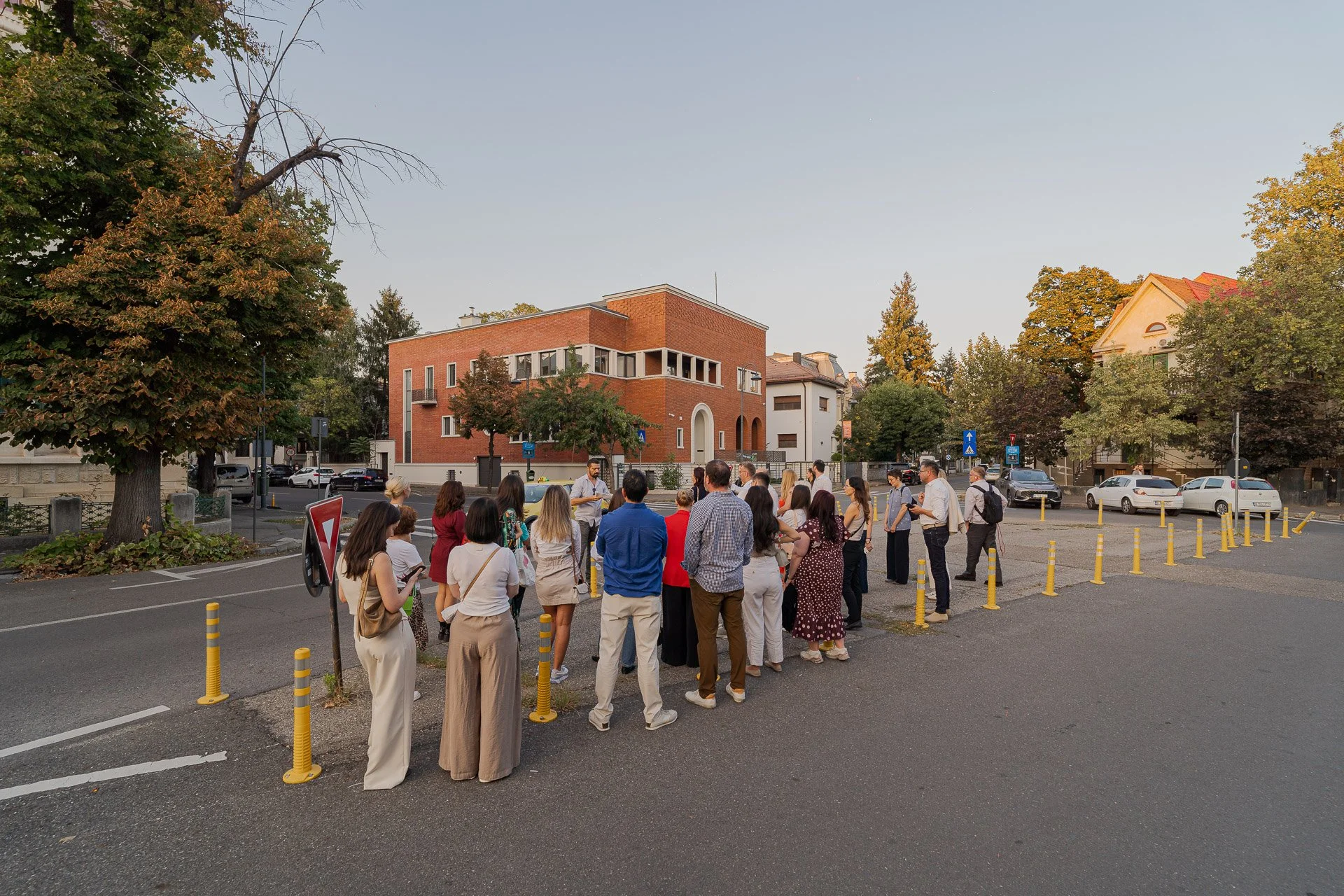 A group of people gathered in an urban outdoor setting during the late afternoon or early evening, with trees, brick and white buildings, and parked cars in the background.