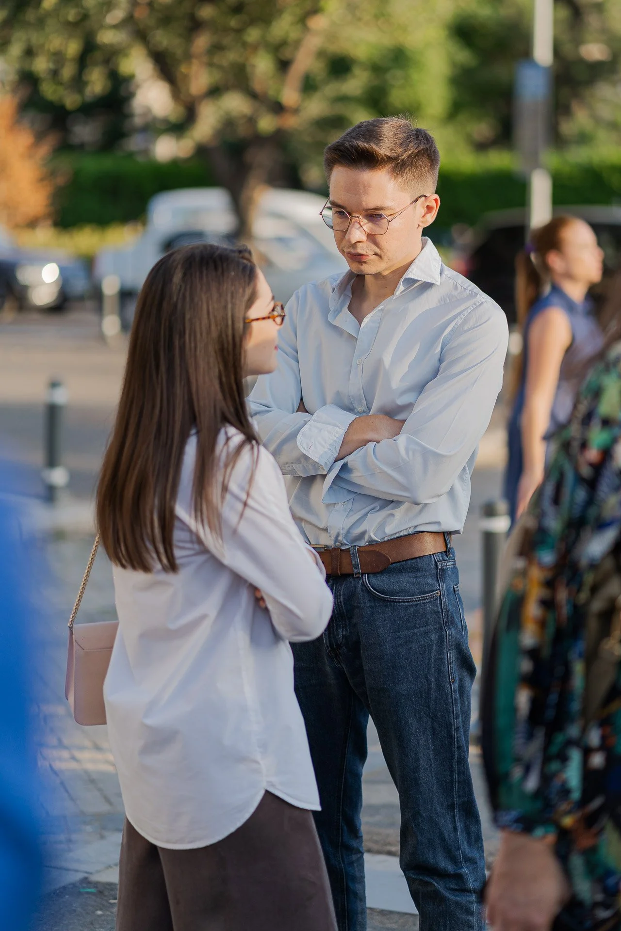 A man with glasses and a woman with glasses talking outdoors among people and trees, with parked cars in the background.