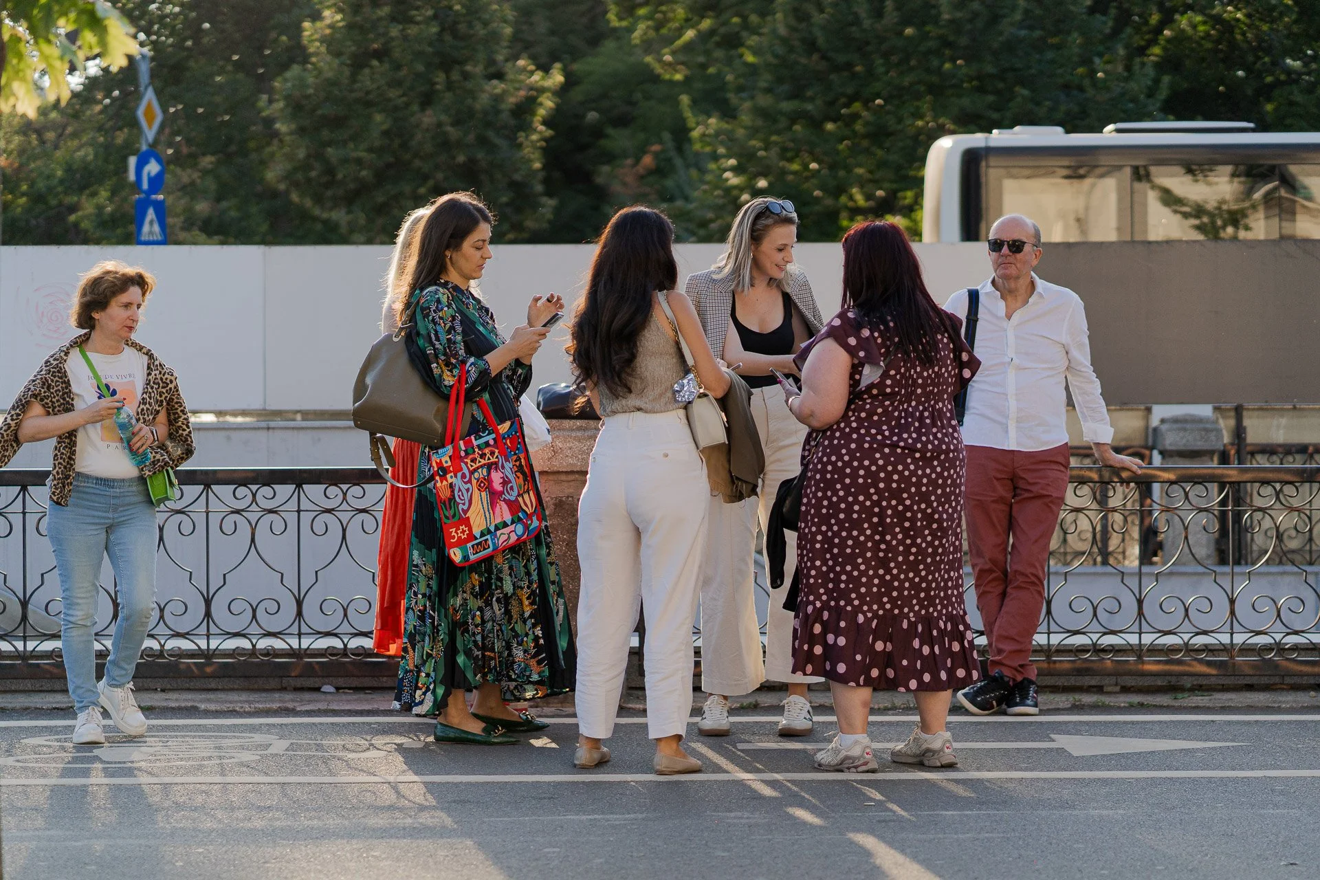 A group of seven diverse people standing outdoors on a sunny day, engaging in conversation, with some looking at their phones. They are near a metal railing and a white wall, with trees and a bus in the background.
