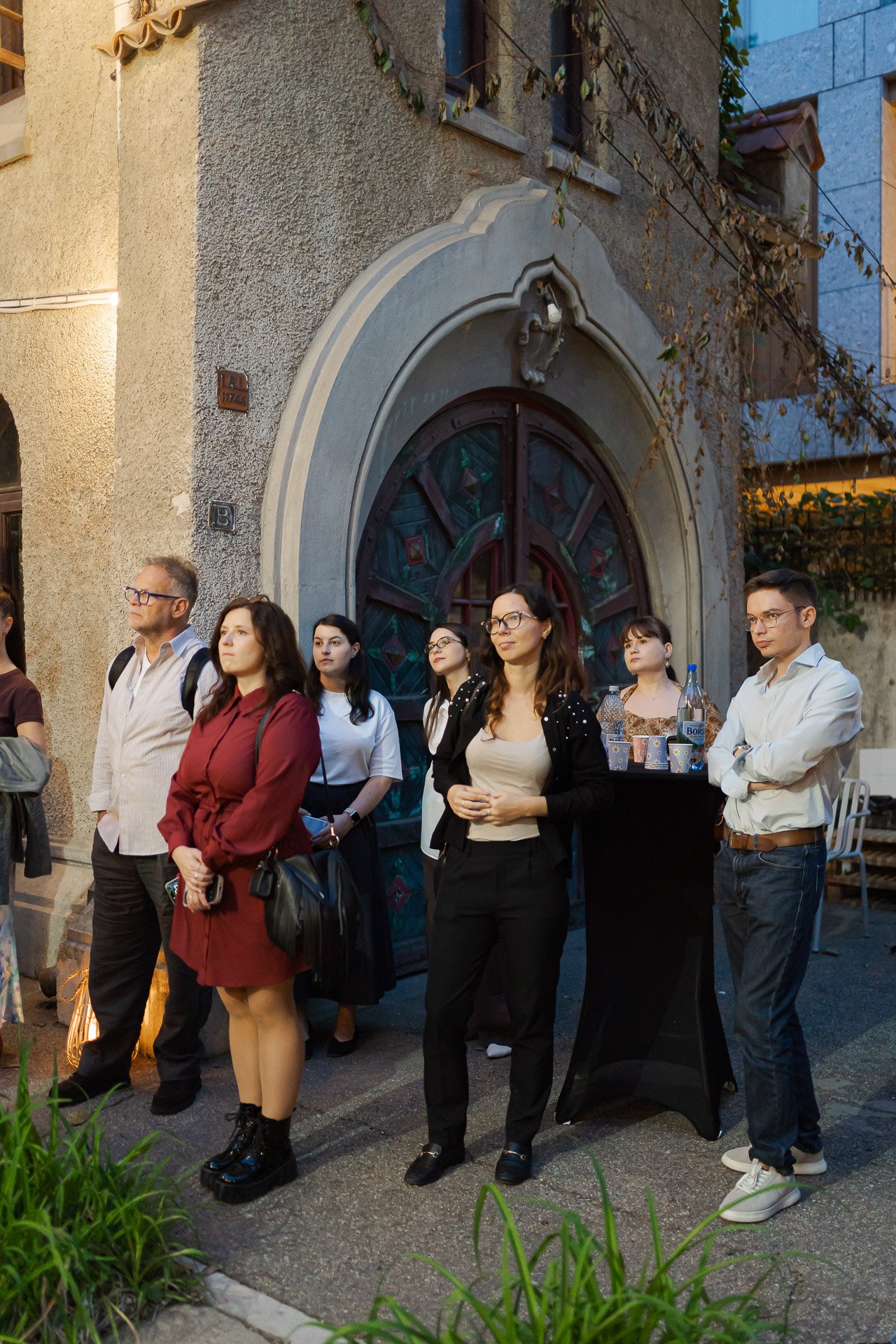 A group of six people standing outside of a building at night, looking attentive. The building has a decorative arched doorway with stained glass windows and a textured exterior wall. Some people hold cups and bottles, and there is a table with bever