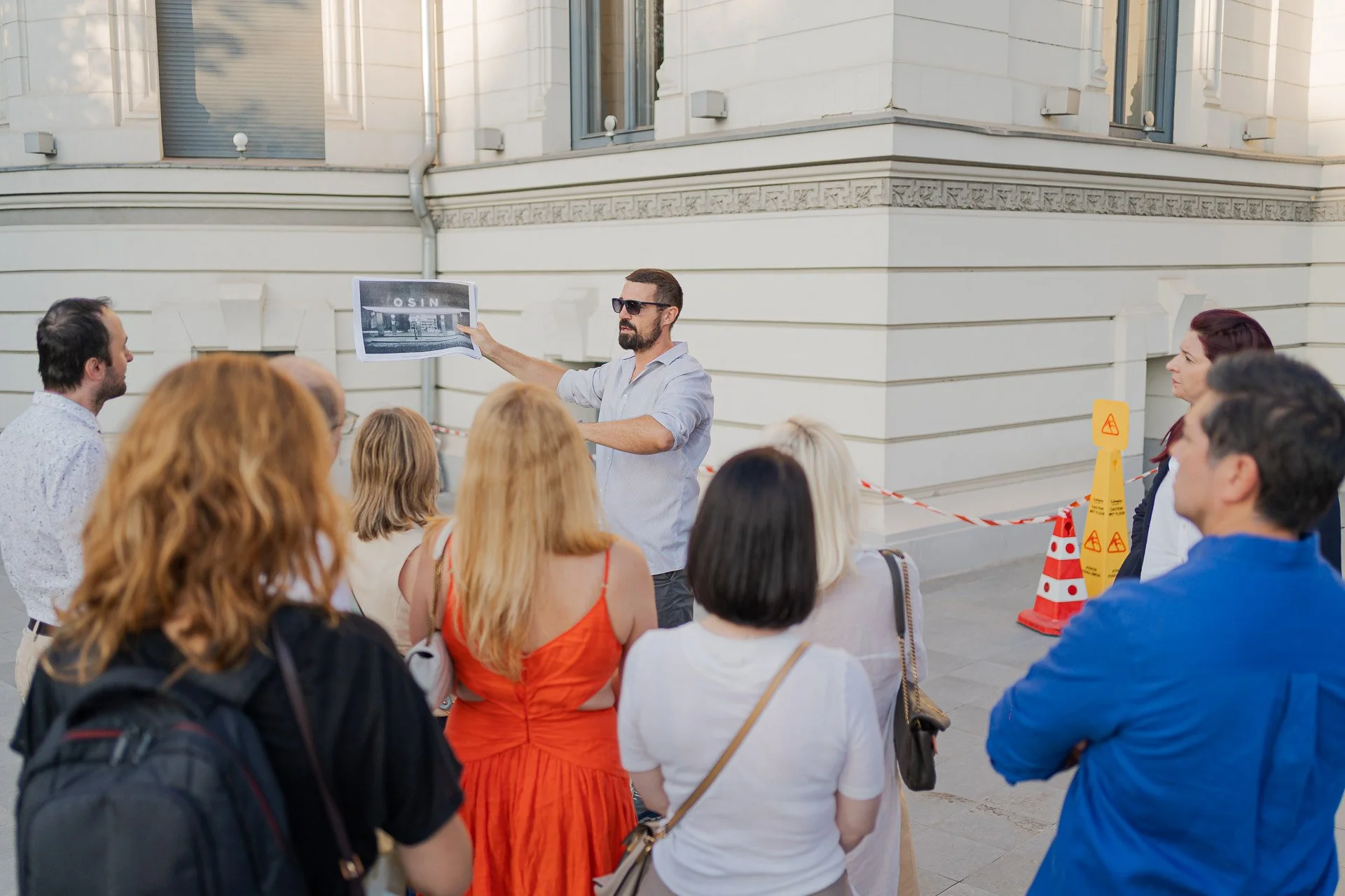A man with sunglasses giving a presentation to a group of people outdoors near a large white building, holding a large black-and-white photograph.