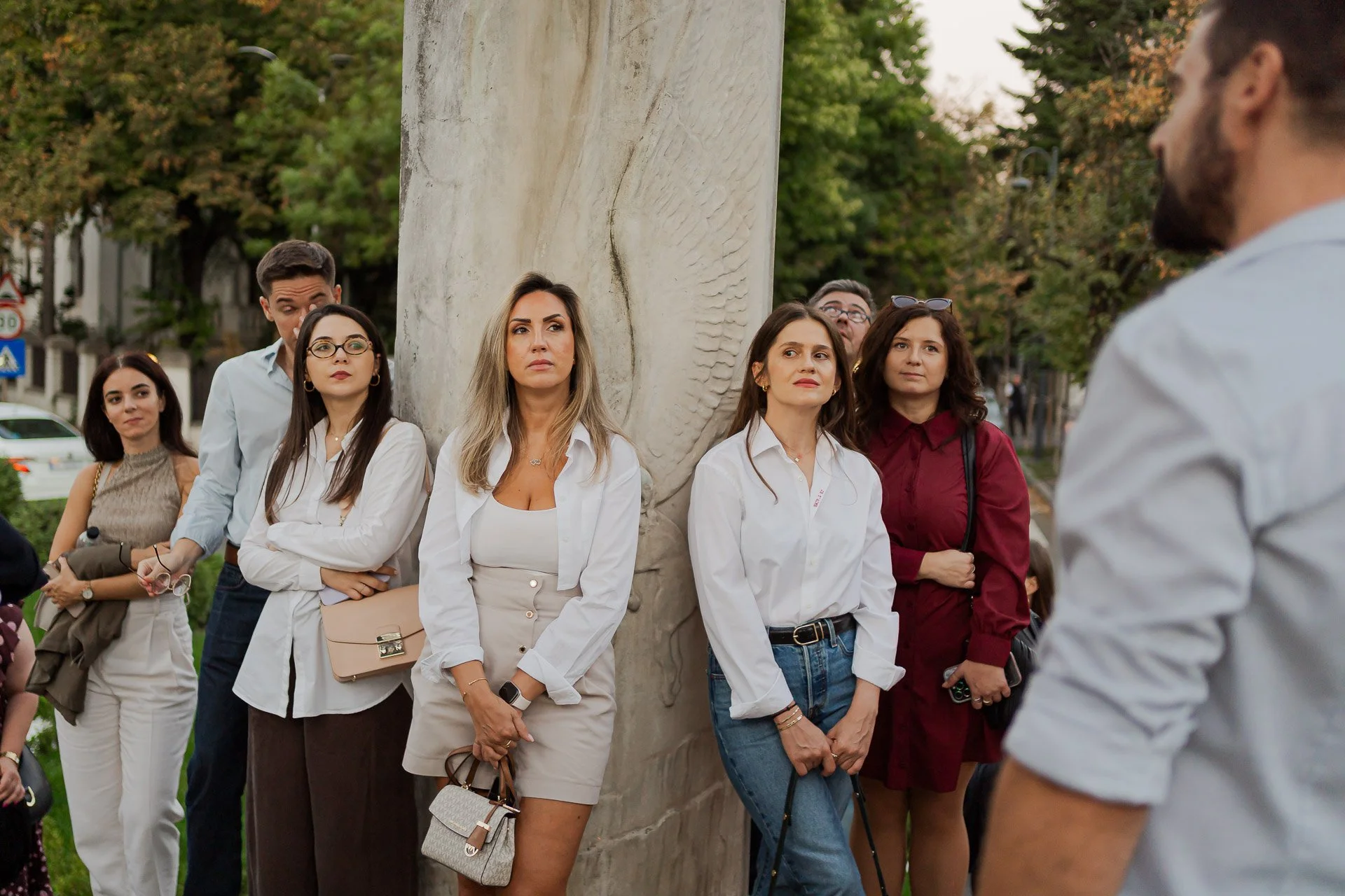 Group of people standing outdoors, listening to a speaker. Some are holding purses, wearing glasses, and dressed in casual to semi-formal attire. Trees with green and autumn-colored leaves are in the background.