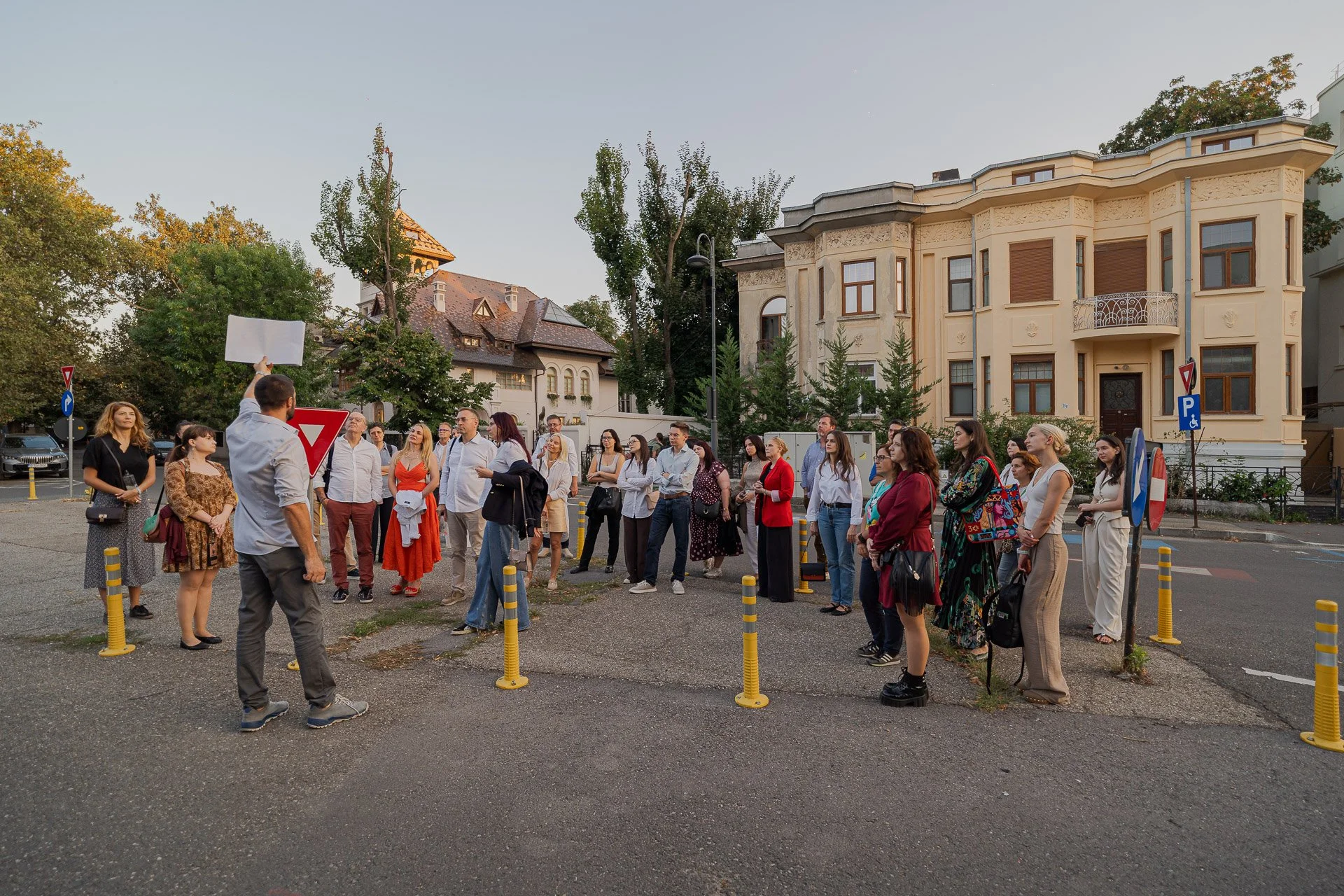 A group of people gather outdoors near a residential street, some listening to a man holding a blank sign, with trees and large houses in the background during evening.