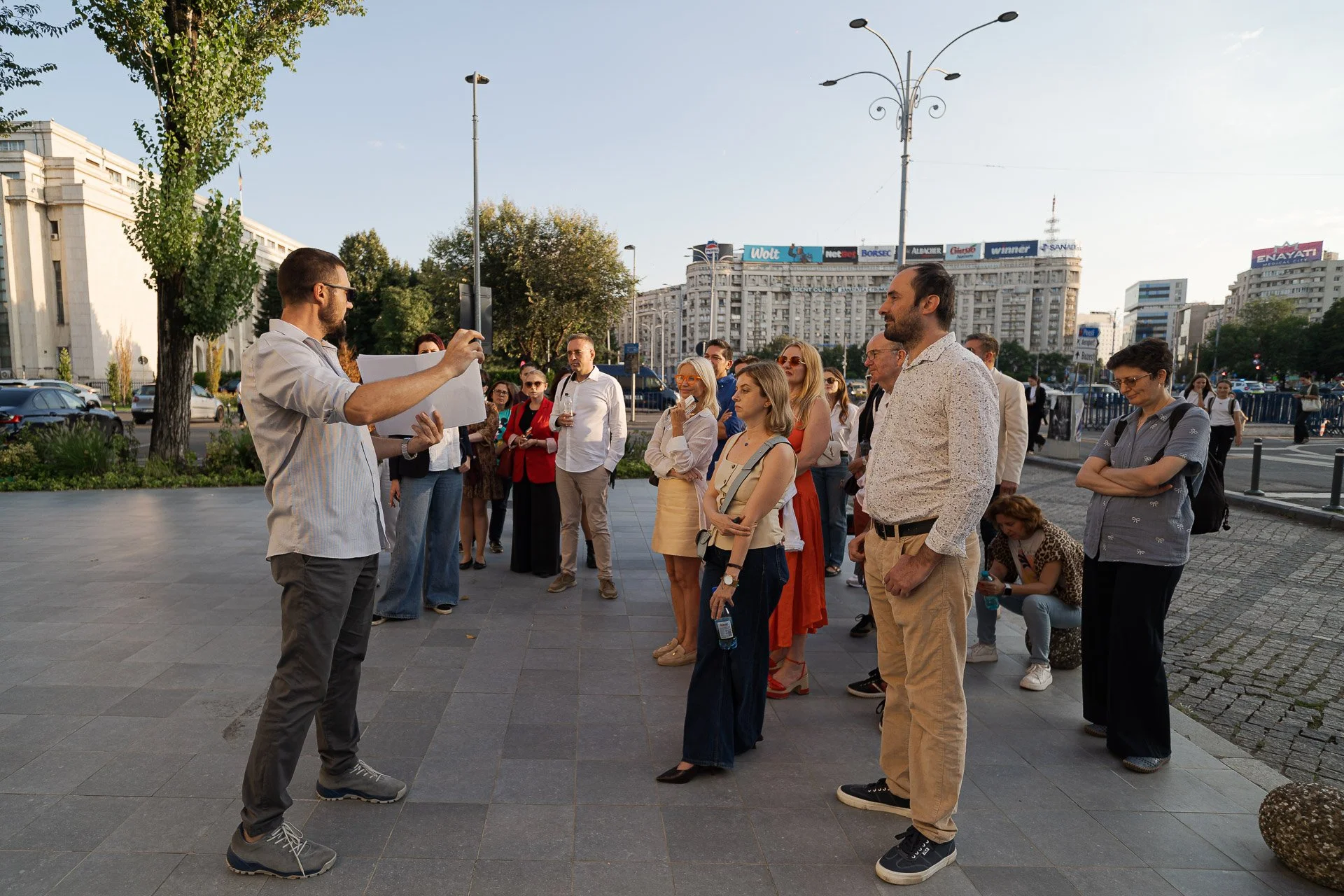 A man holding a phone is speaking to a group of people gathered outdoors in an urban setting, with buildings, trees, and cars in the background.