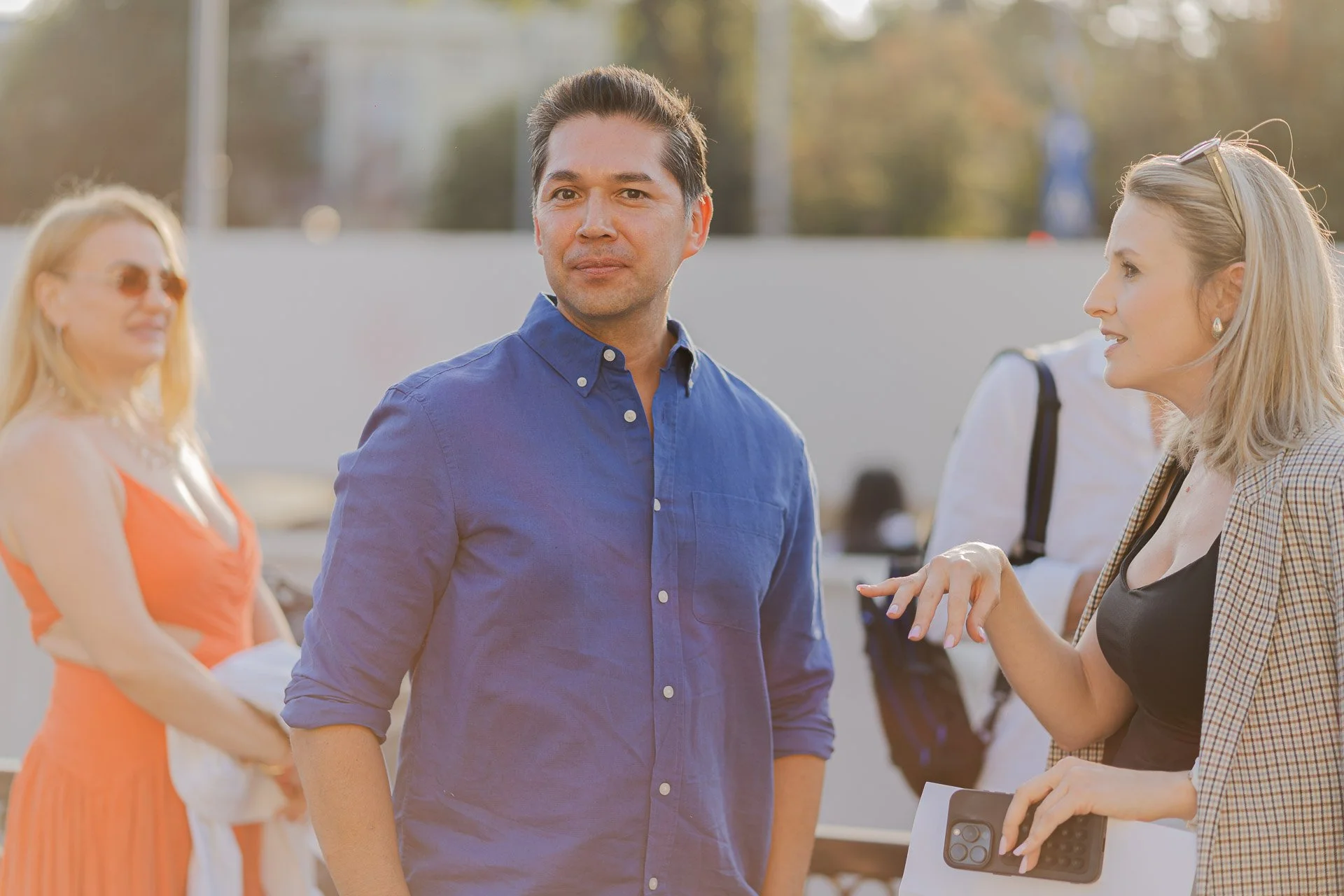 A group of people outdoors, with a man in a blue shirt at the center, talking to a woman holding a clipboard and smartphone.