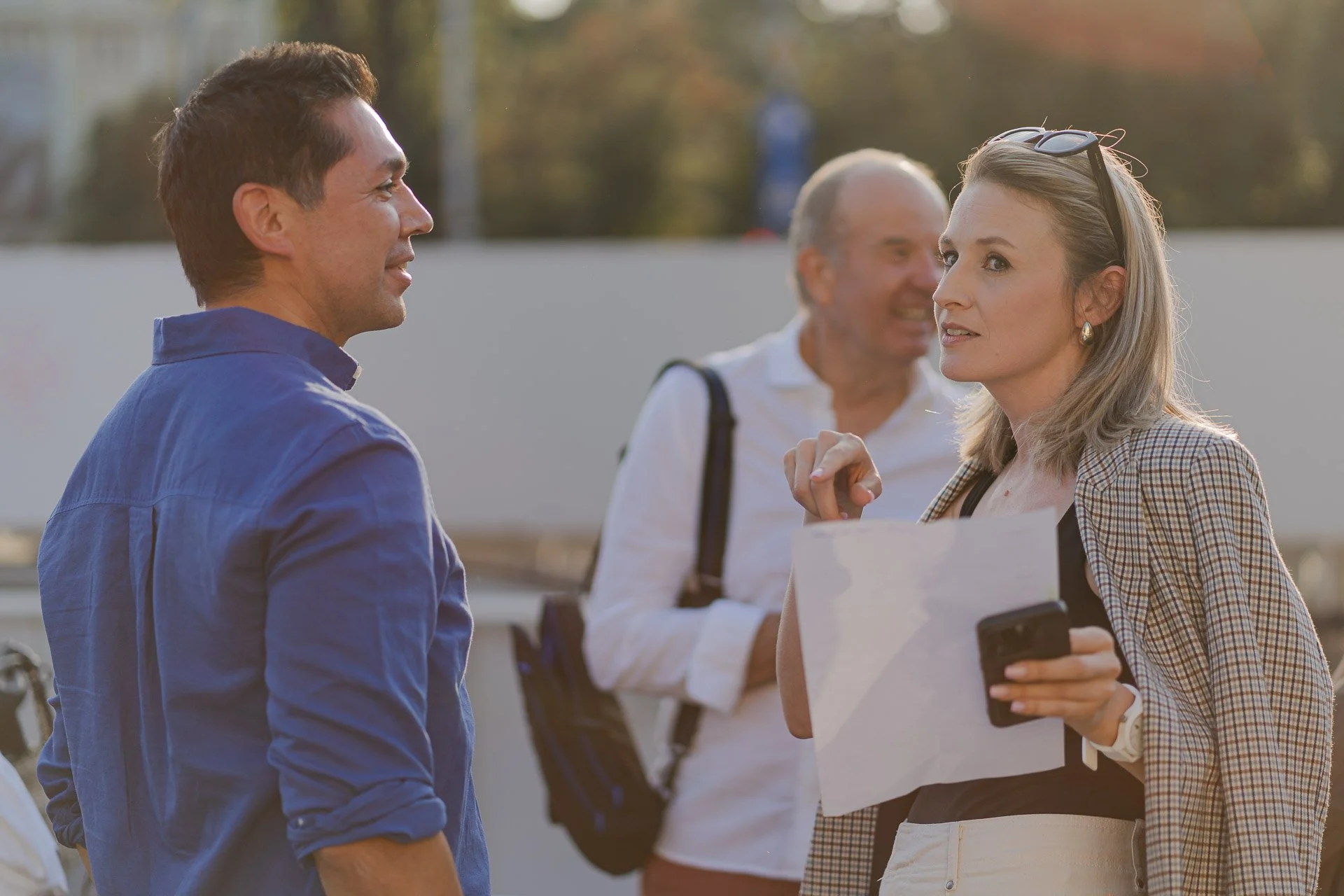 A man and a woman are having a conversation outdoors, with a woman in the background holding a smartphone and a paper. The woman in the foreground is wearing glasses on her head and earrings, and the man is dressed in a blue shirt. They are smiling a