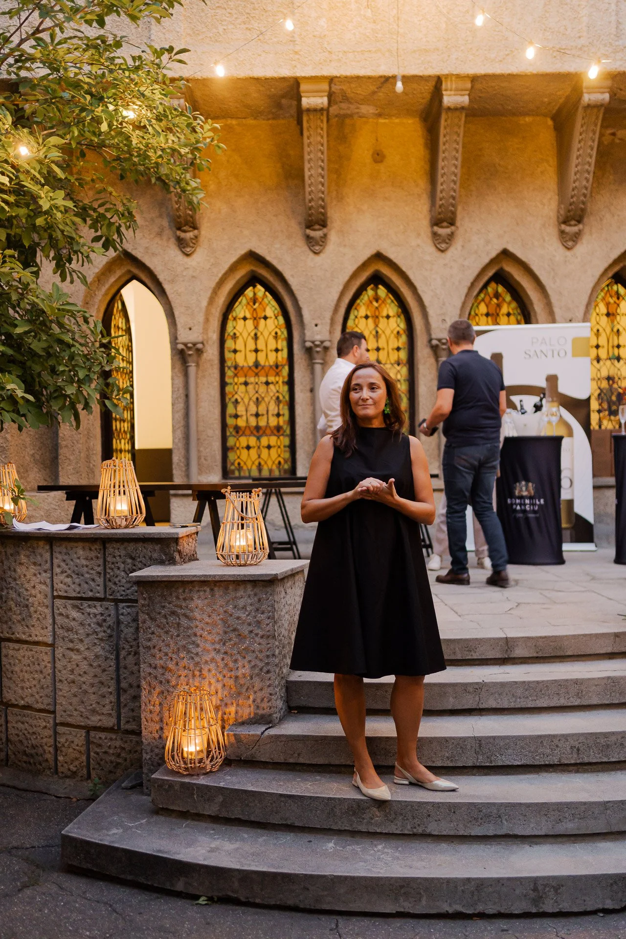A woman in a black dress standing on stone steps outdoors at an evening event, illuminated by candle lanterns, with a historic building with stained glass windows and string lights in the background.