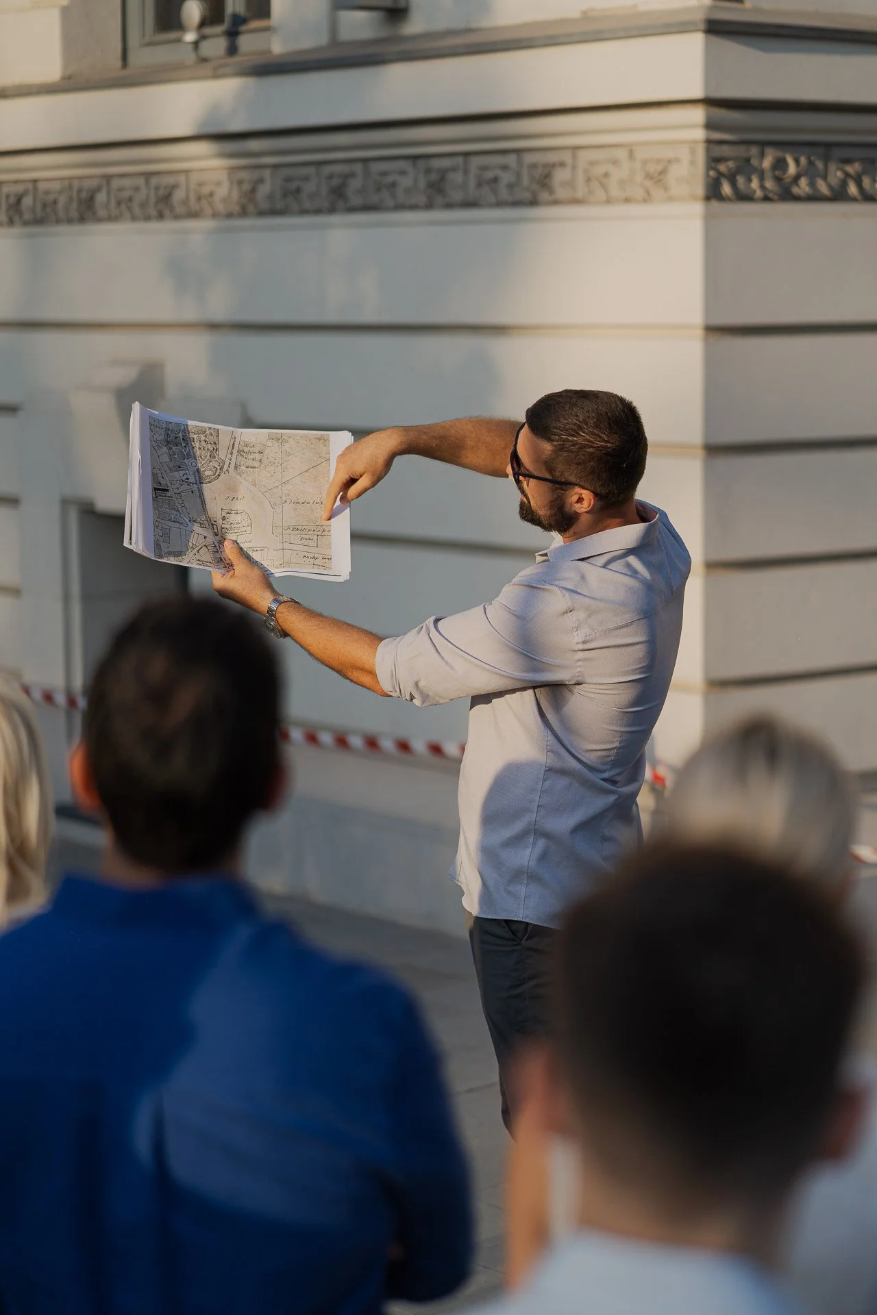 Man in glasses holding and explaining a large paper map to a group of people outdoors.