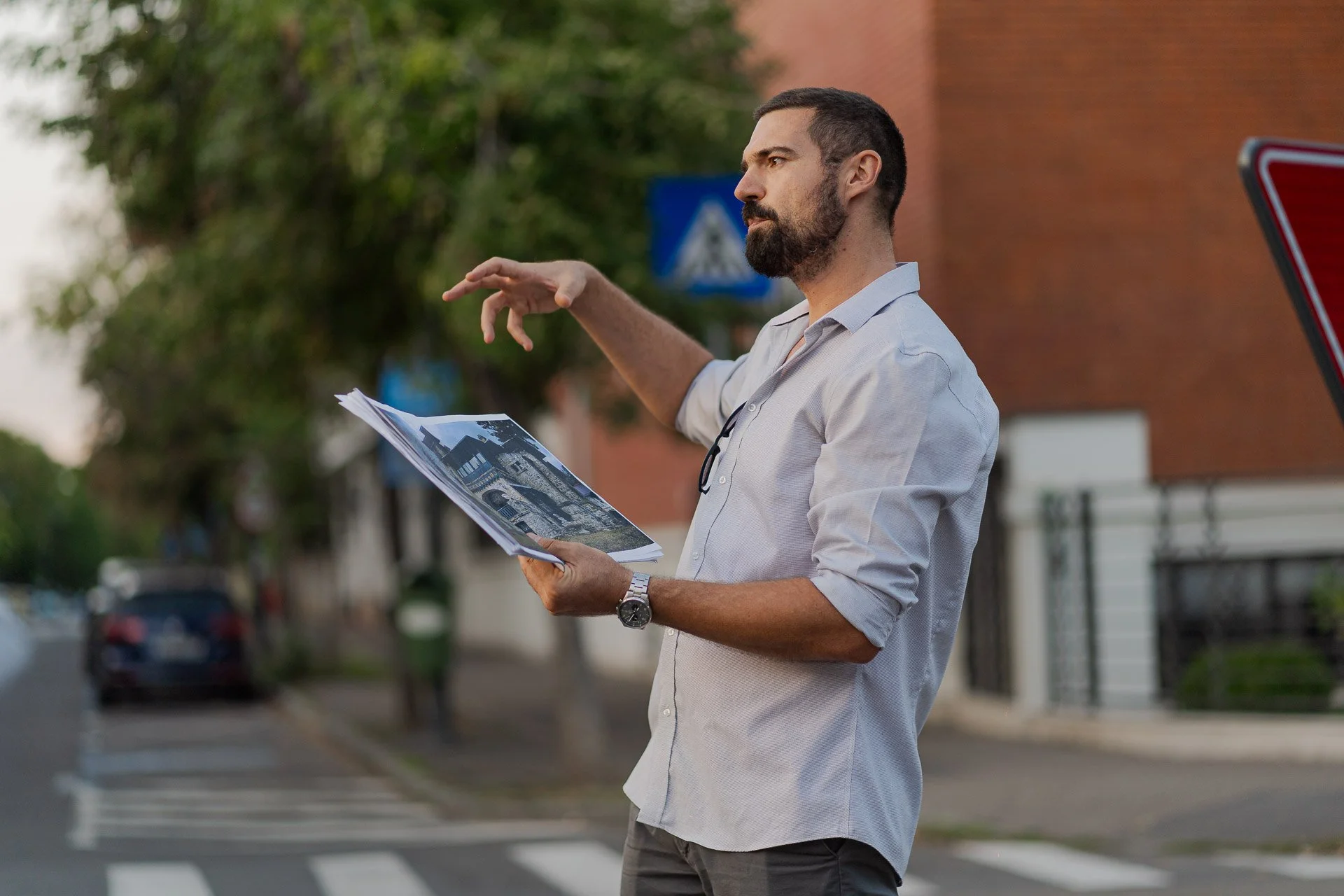 A man with a beard and dark hair, wearing a white dress shirt, holding papers with building images, standing near a crosswalk on a city street, gesturing with his left hand.