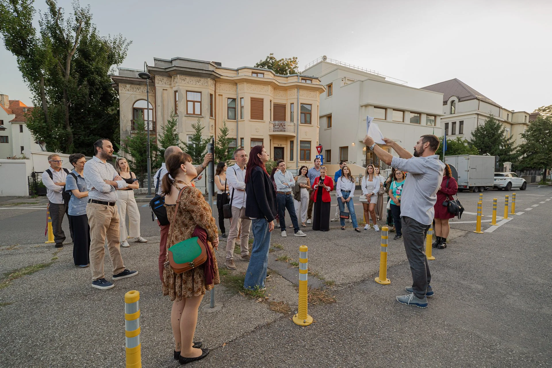 A group of people participating in an outdoor guided tour or rehearsal in a parking lot, with a man leading and holding papers, surrounded by modern residential buildings and trees.