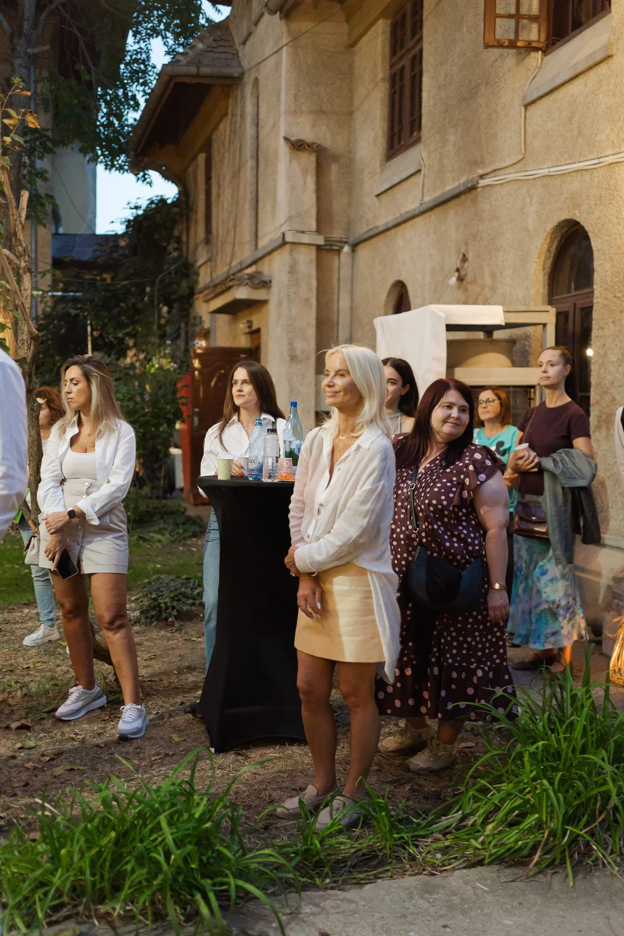 Group of women gathered outdoors near an old brick building during the evening. Some women are dressed in casual and semi-formal attire, standing around a small table with drinks, engaged in conversation and observing something outside the frame.