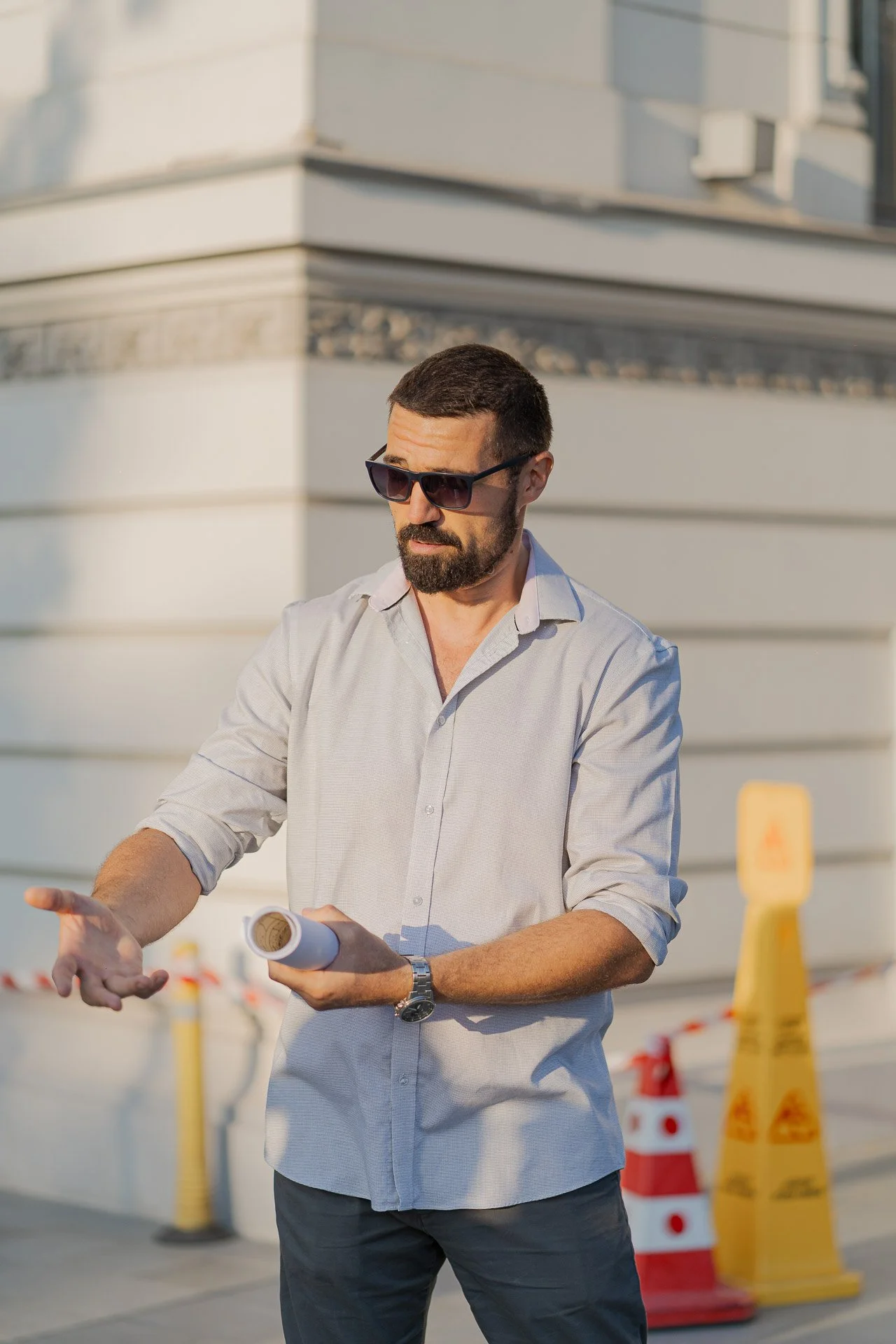 Man with dark hair and beard wearing sunglasses, a light gray button-up shirt, and a watch, holding a rolled-up document or blueprint, standing outdoors near construction cones and barriers, with a building in the background.