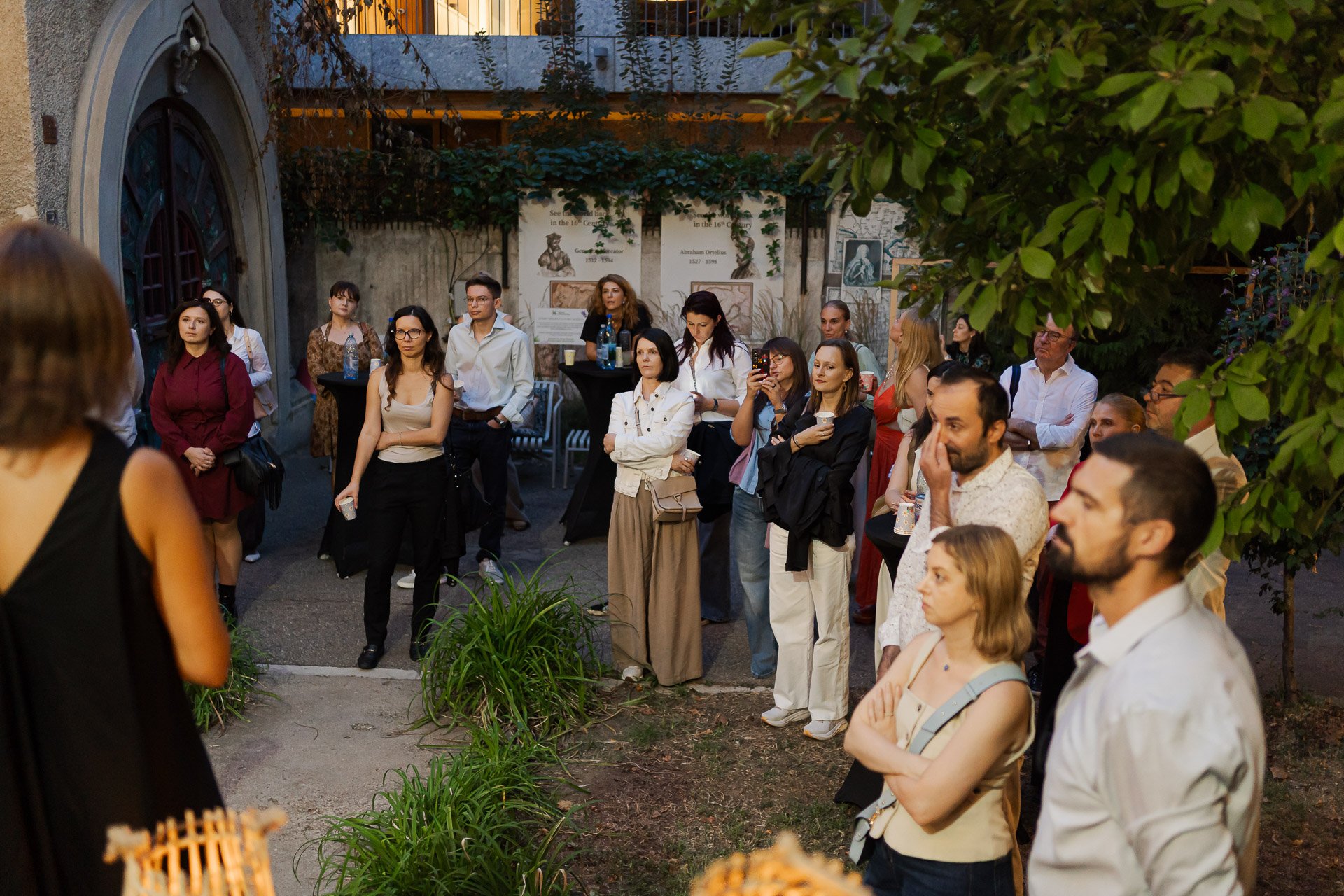A group of people gathered outdoors during the evening, listening attentively to a speaker. The crowd consists of men and women standing and observing, with some individuals taking photos or holding drinks. The scene includes lush greenery and a buil