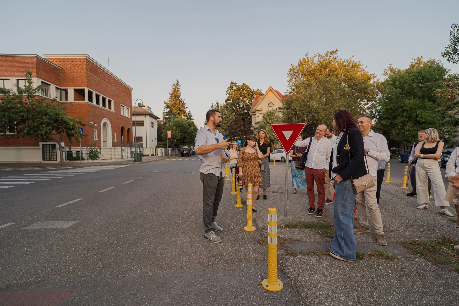 Group of people standing by a yield sign on a city street during the evening with trees and buildings in the background.