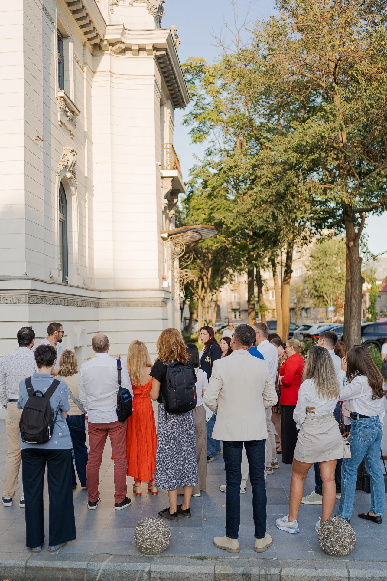 Group of people standing on a sidewalk in front of a large white historic building, with trees and parked cars in the background, during daytime.
