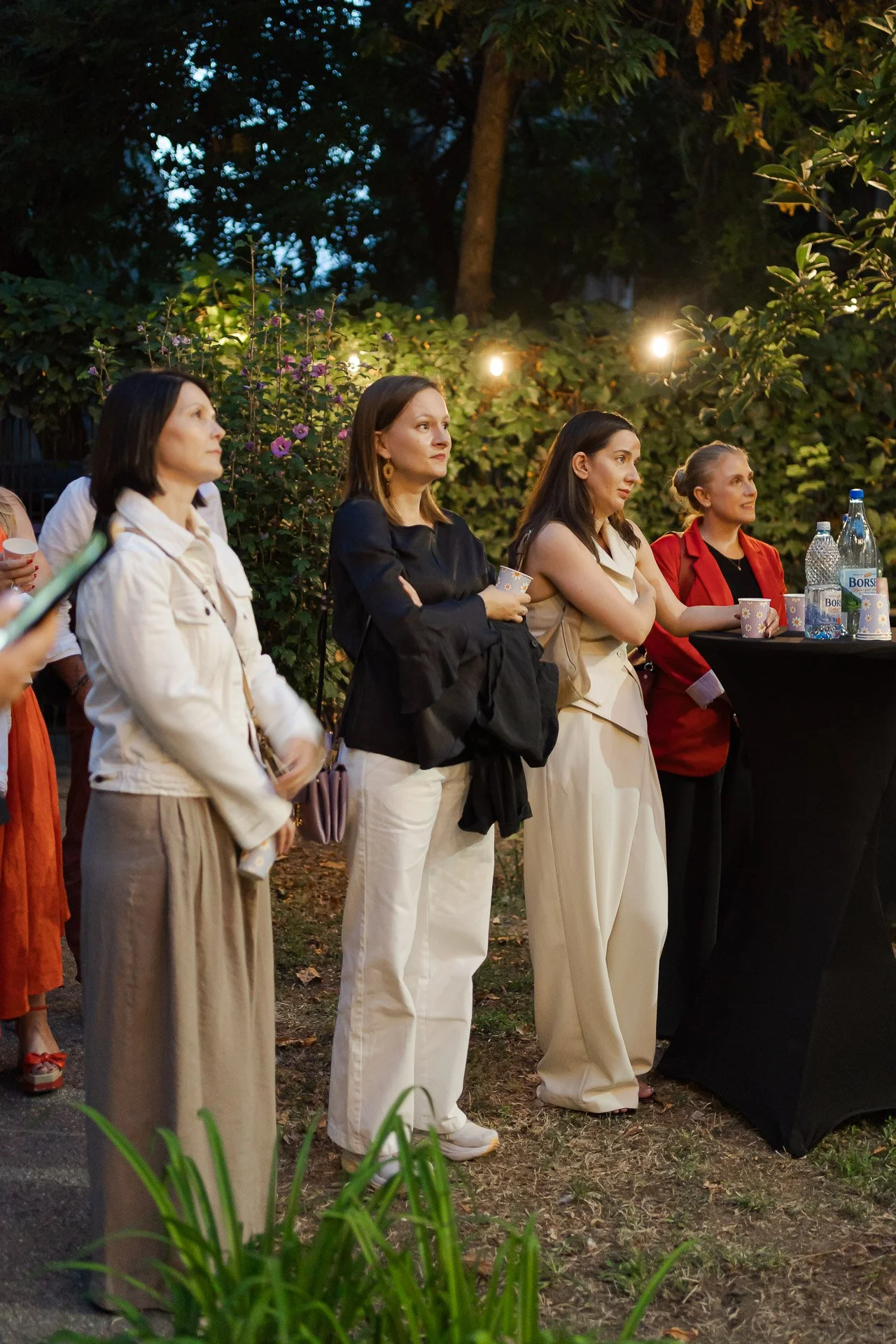 Group of women standing outdoors in the evening with greenery and string lights in the background, some holding cups and looking attentively at something.