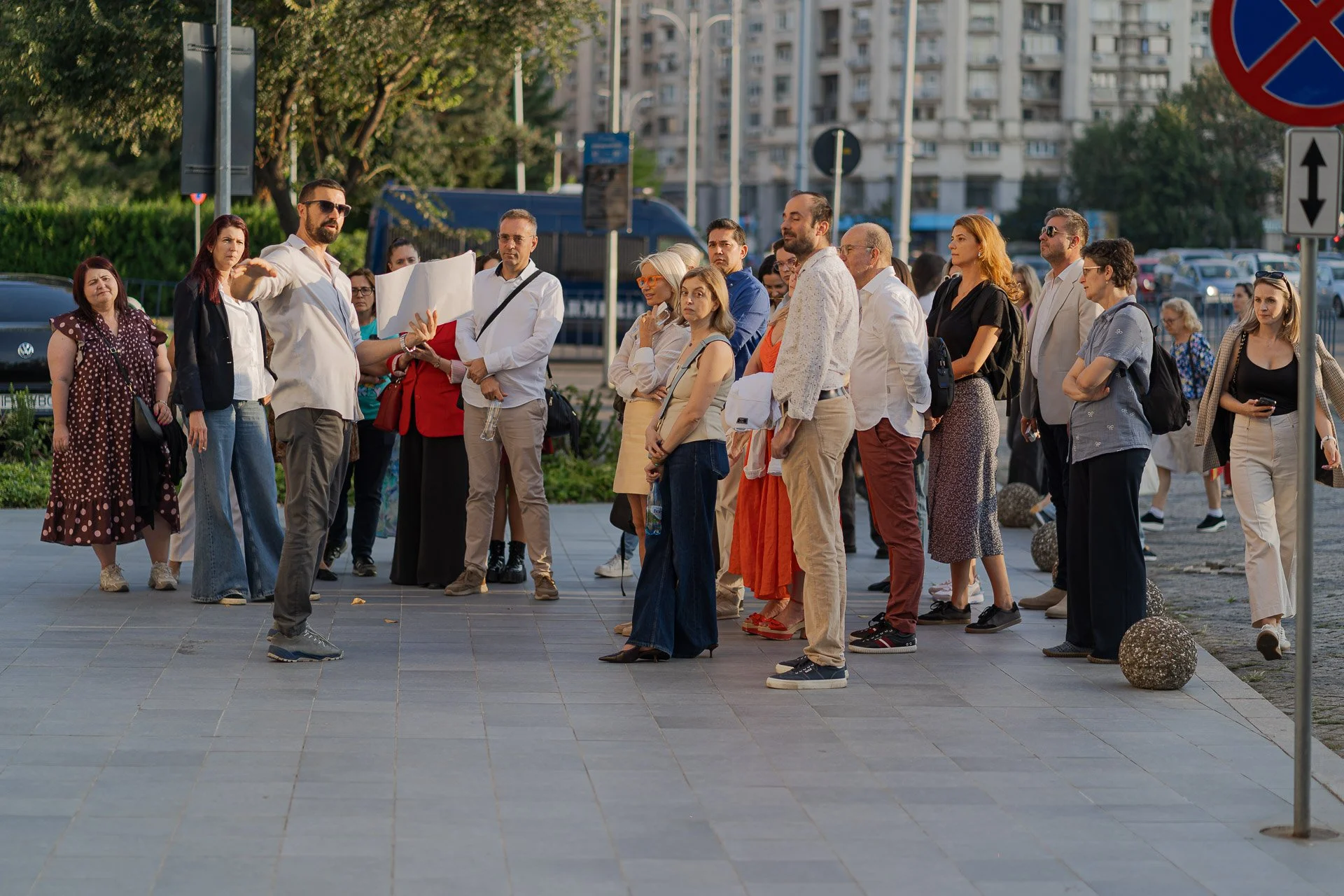 A group of people standing in a line outdoors in an urban area, with some individuals engaged in conversation and others looking around, surrounded by trees, buildings, and street signs.