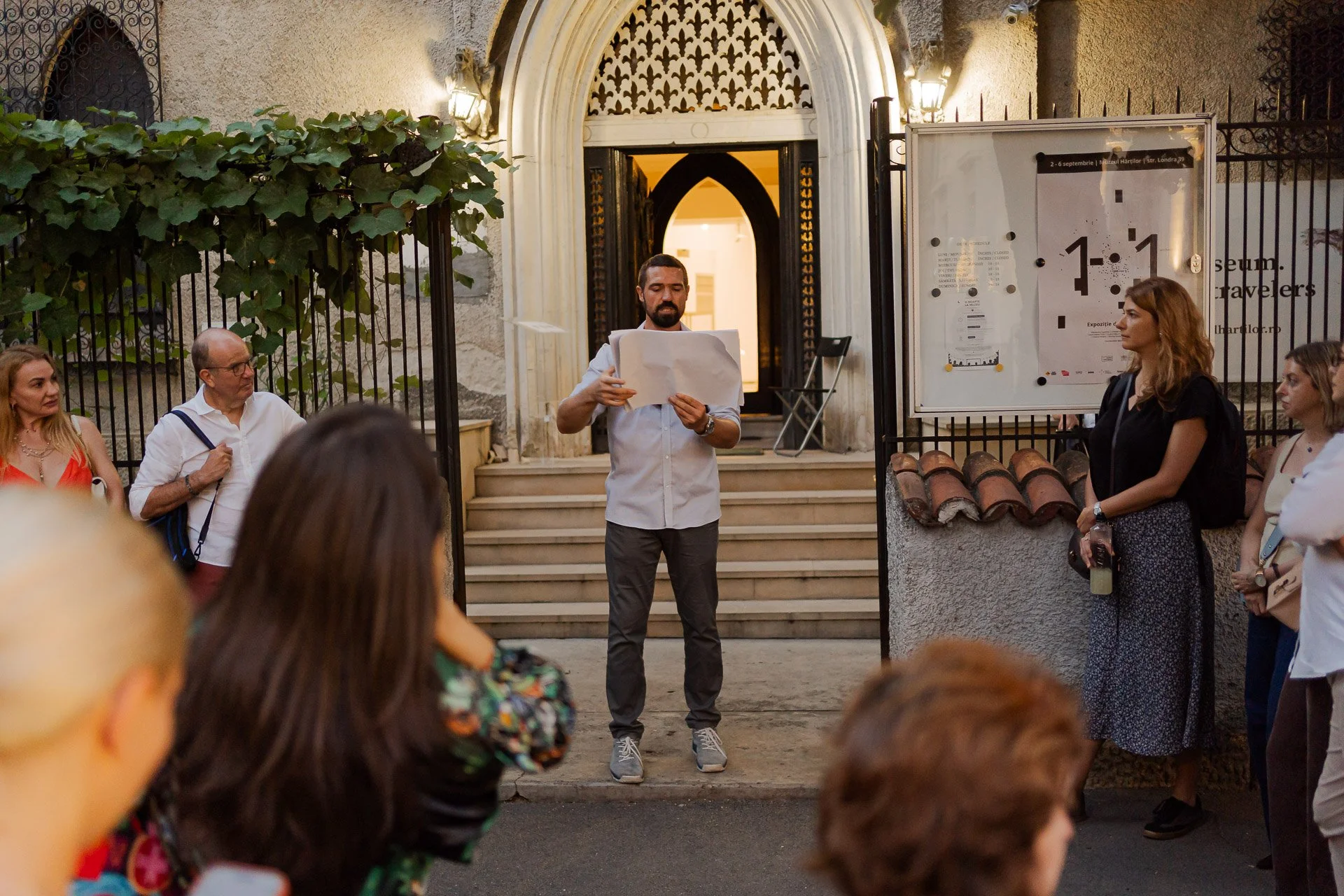 A man with a beard and gray pants reads from a paper at an outdoor gathering in front of an arched doorway, surrounded by people listening.