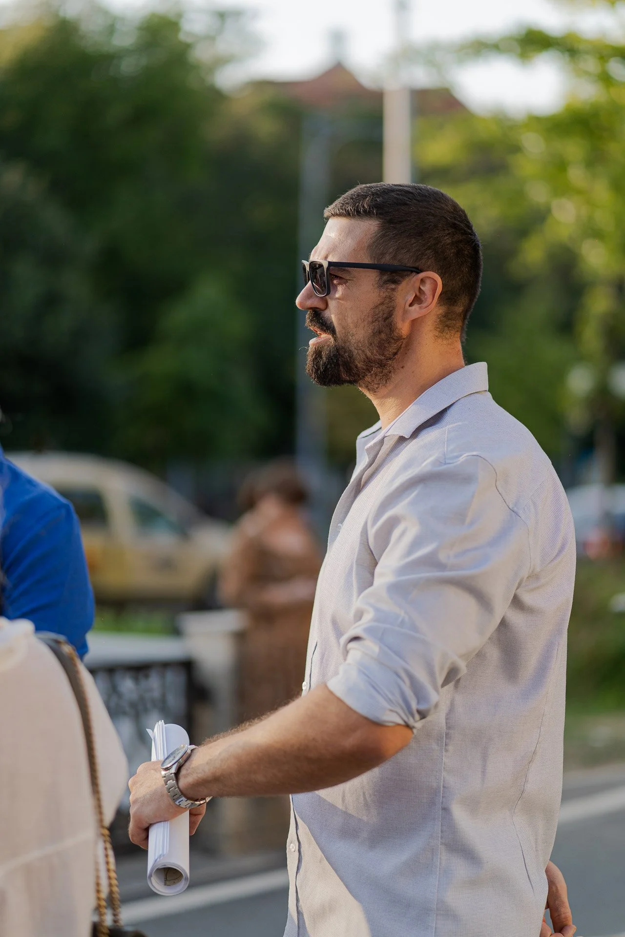 Profile of a man with short dark hair, beard, sunglasses, wearing a light-colored button-up shirt, holding rolled papers, outdoors with blurred trees and vehicles in the background.