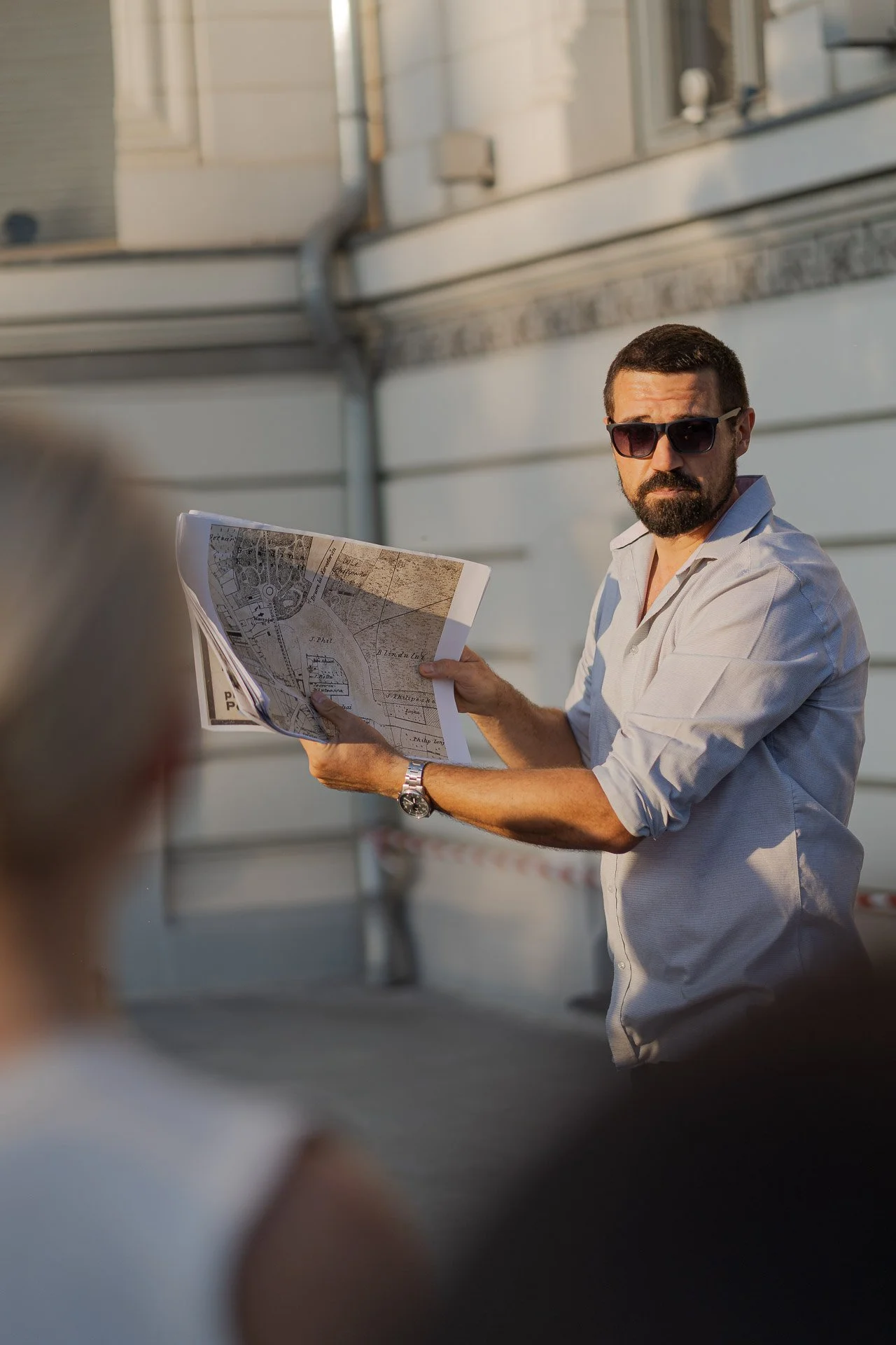 A man wearing sunglasses and a light-colored shirt is holding and looking at a city map or blueprint outdoors, with a building in the background.