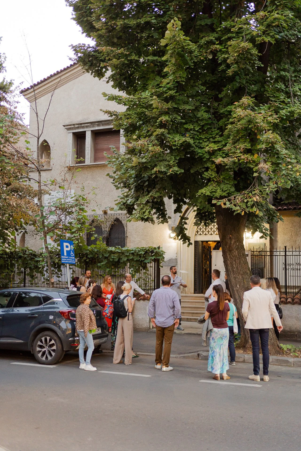People gathered outside a building with a large tree in front, near parked cars and a designated parking sign.