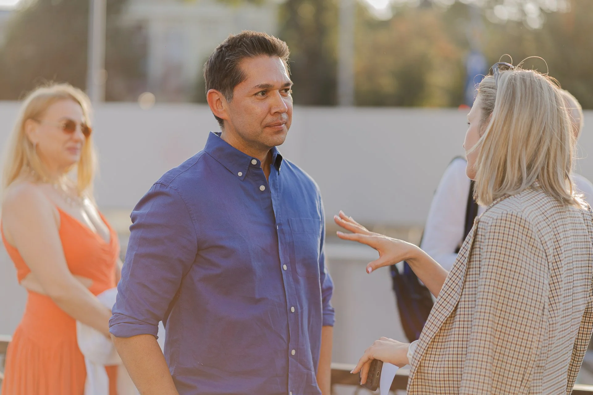 A woman and a man are having a conversation outdoors, with the woman gesturing and speaking to the man. There are other women in the background, one wearing an orange dress and sunglasses, and trees are visible in the distance.