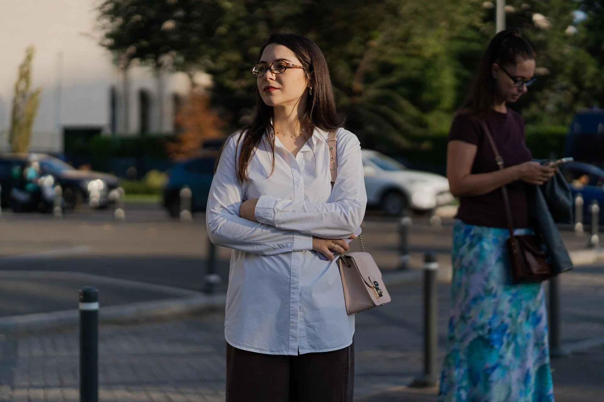 A woman with dark hair and glasses wearing a white blouse, standing outdoors with her arms crossed, during daytime, with another woman in the background looking at her phone.
