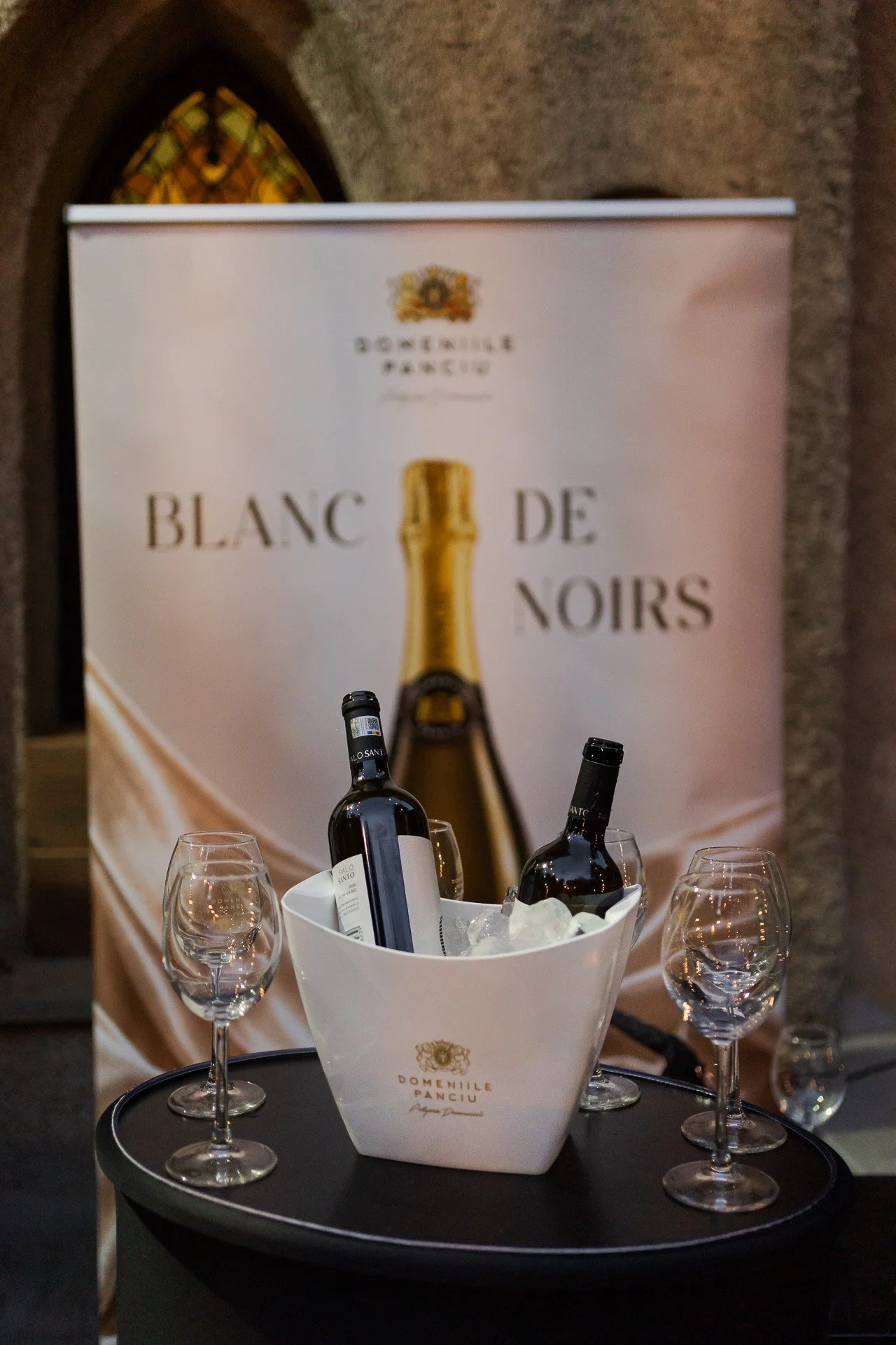 A table with champagne bottles chilling in a white bucket and four empty wine glasses. A large promotional banner in the background features a champagne bottle and the words 'Blanc de Noirs' and 'Domaine Pancyu'.