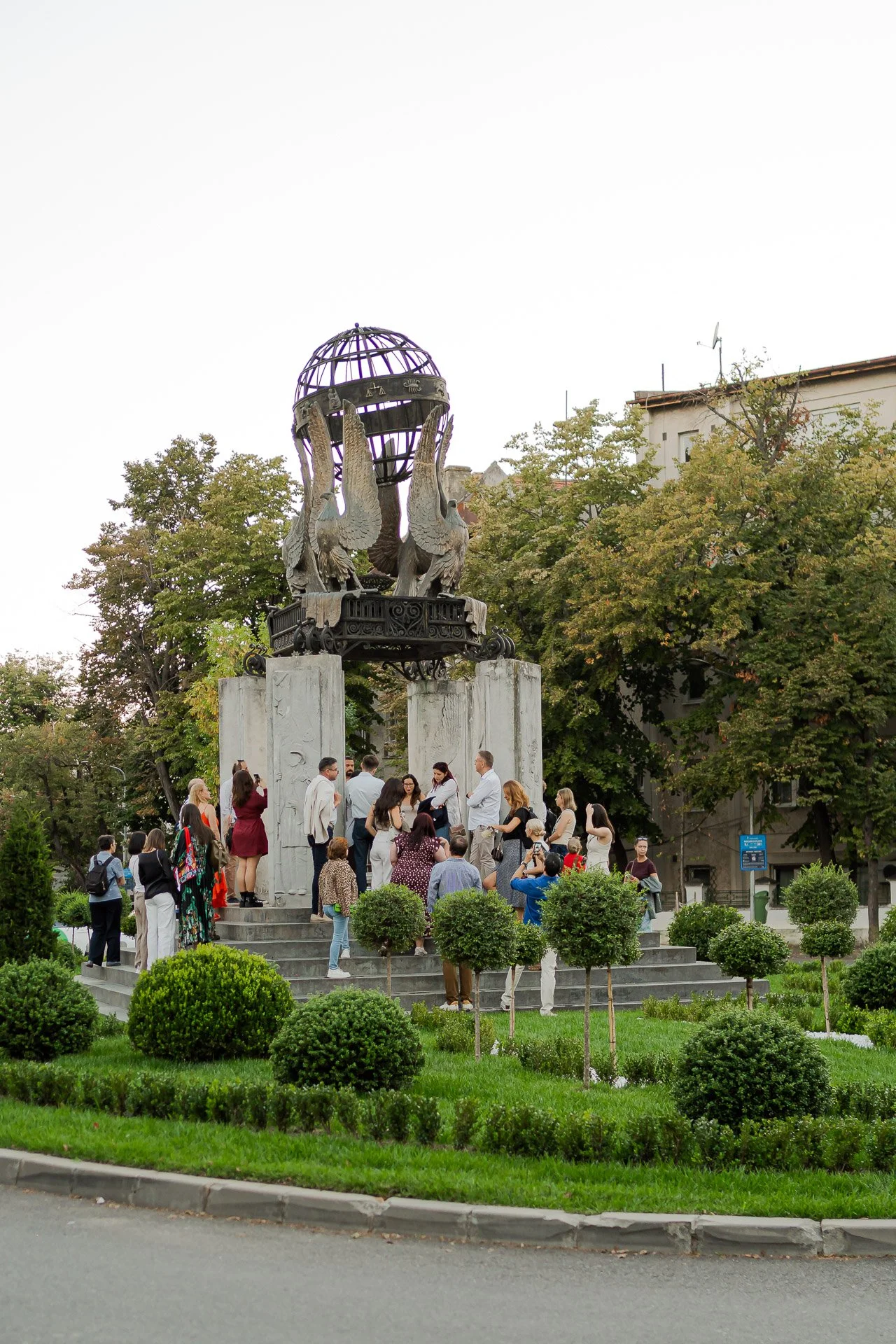 A group of people gathered around a monument in a park with trees and buildings in the background.