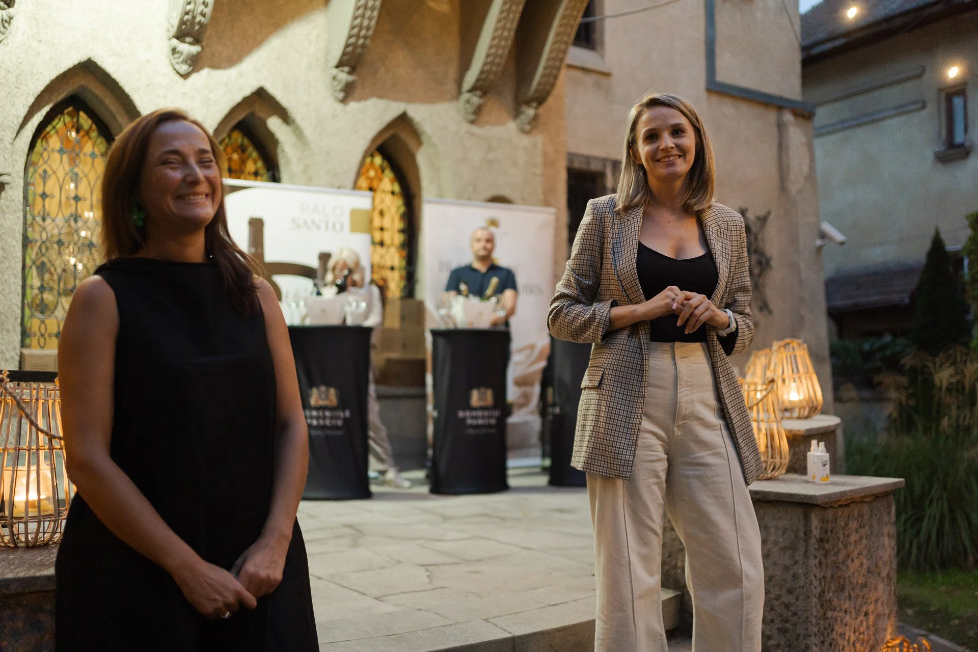 Two women standing outdoors near a building at dusk, smiling and looking at the camera. There are tables and banners in the background.