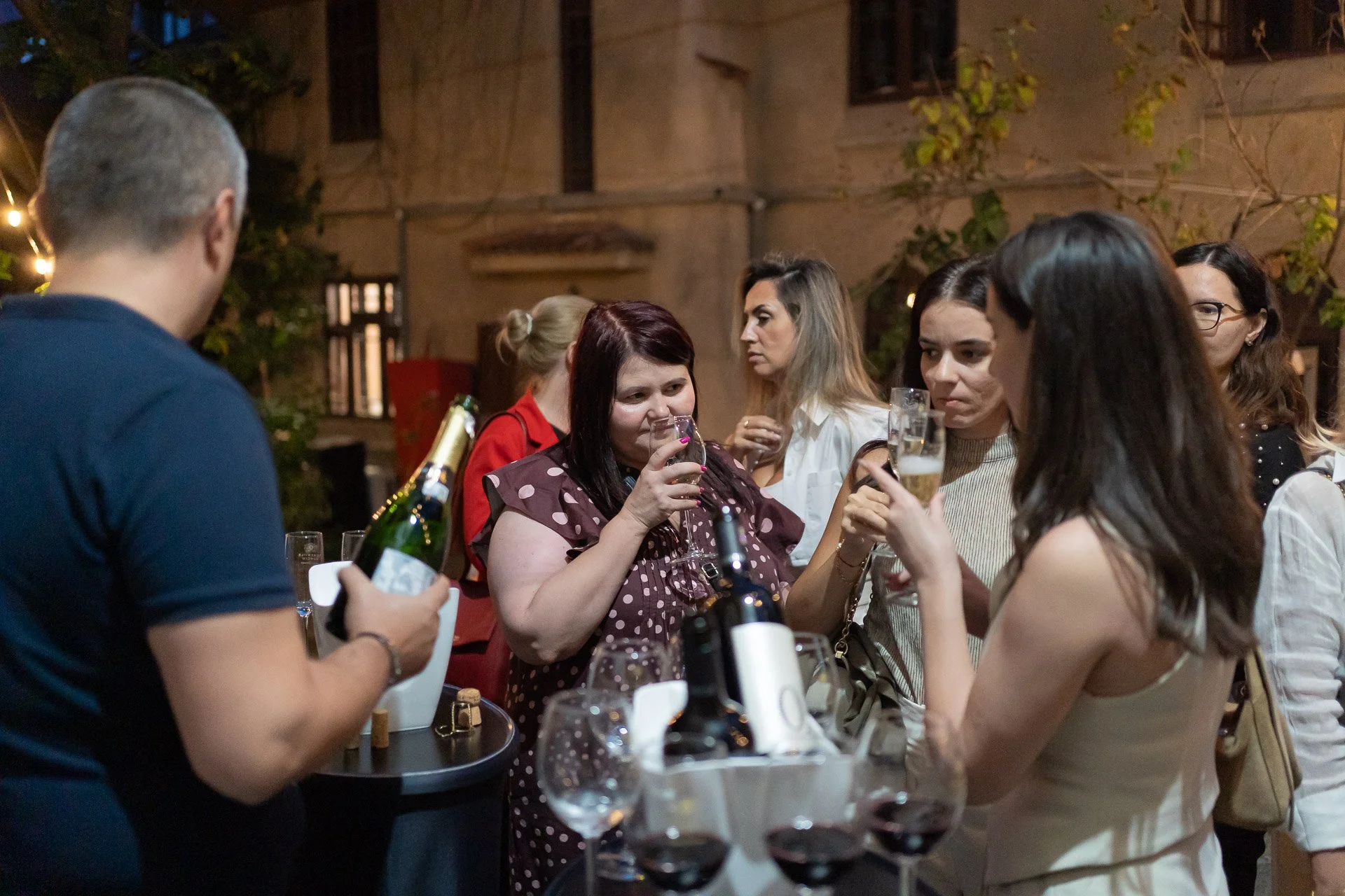 A group of people socializing at a party or gathering; some are holding drinks, including wine glasses and beer bottles, in an outdoor setting with warm lighting and a building in the background.