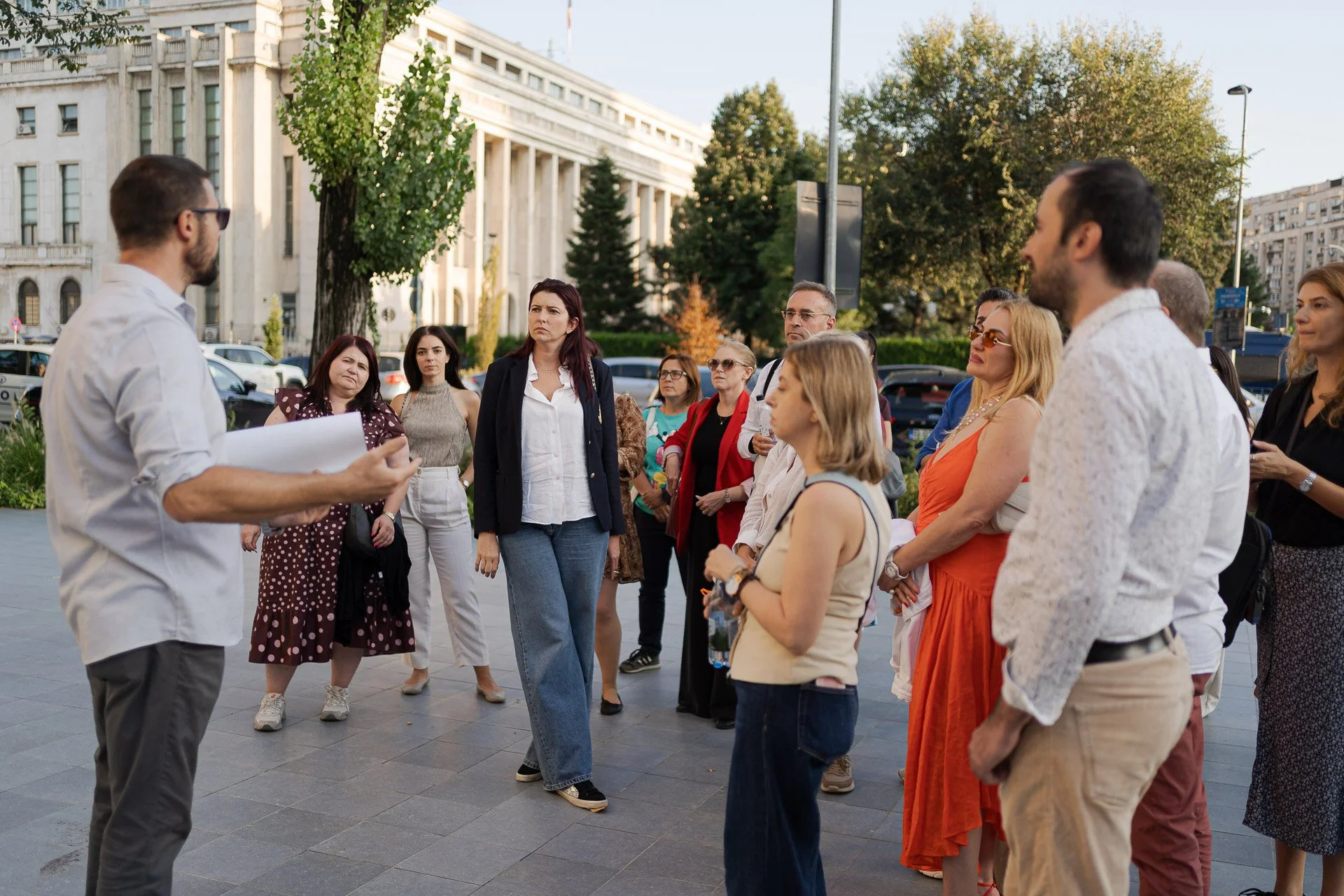 A man with glasses and a white shirt speaks to a group of diverse people outdoors near a large building with trees in the background. The group listens attentively.