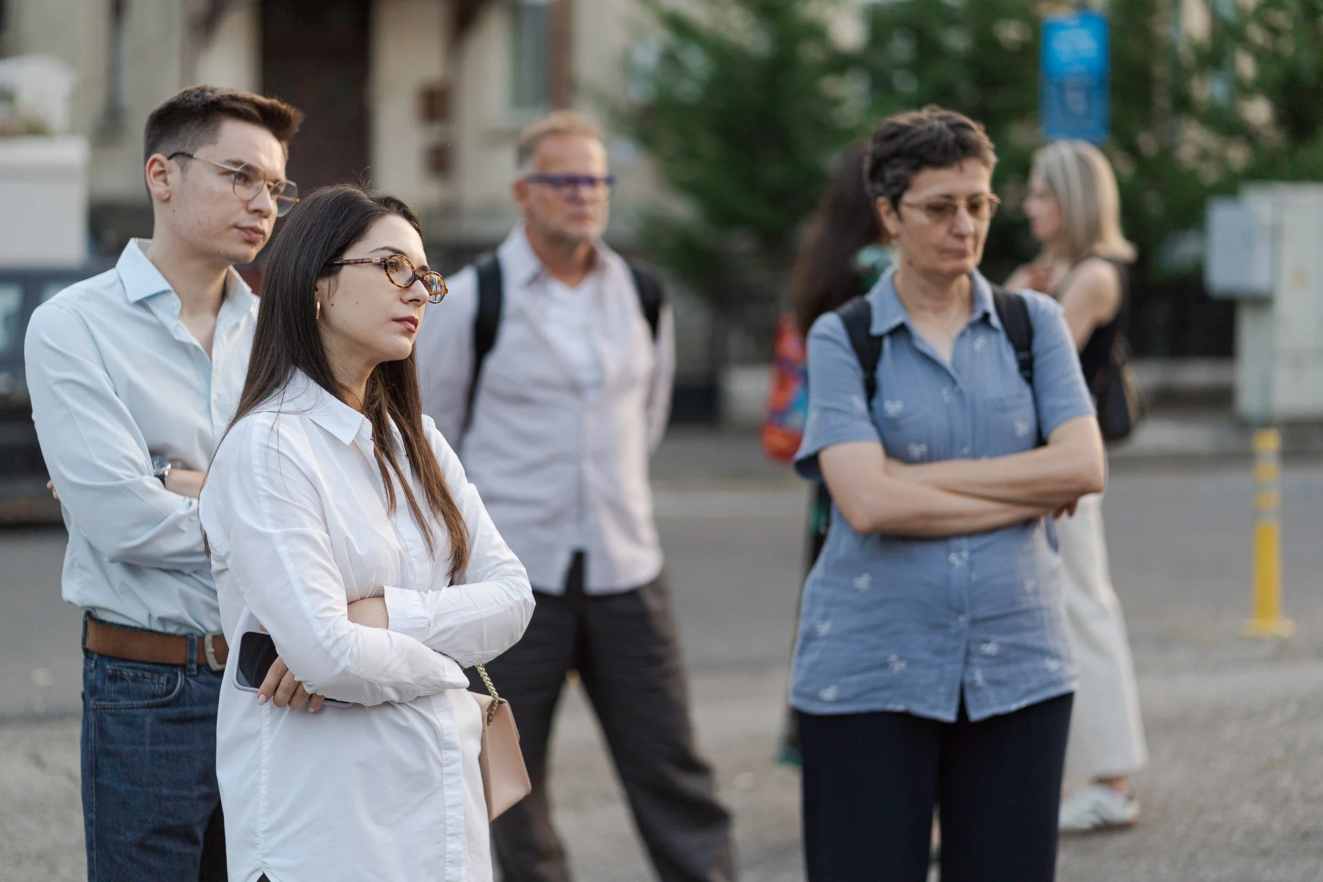 A group of five adults standing outdoors, appearing contemplative or serious, with arms crossed or hands at their sides, in front of a blurred background of a street and trees.