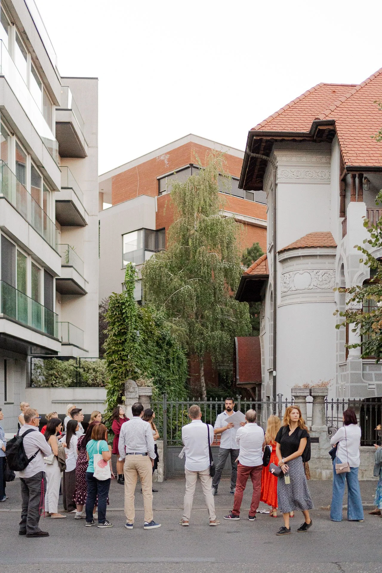 A group of people gathered in front of a historic building with white walls, decorative architectural elements, and a red-tiled roof, while modern apartment buildings are visible in the background.