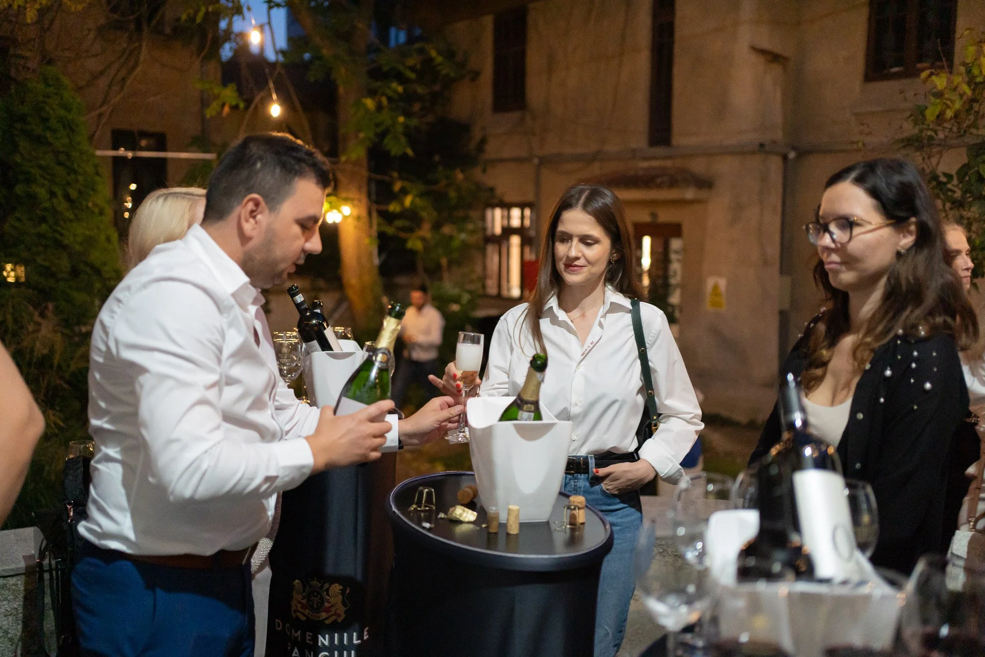 People at an outdoor evening party with drinks and wine bottles, with a man pouring and a woman holding a champagne glass, in a cozy, dimly lit setting.