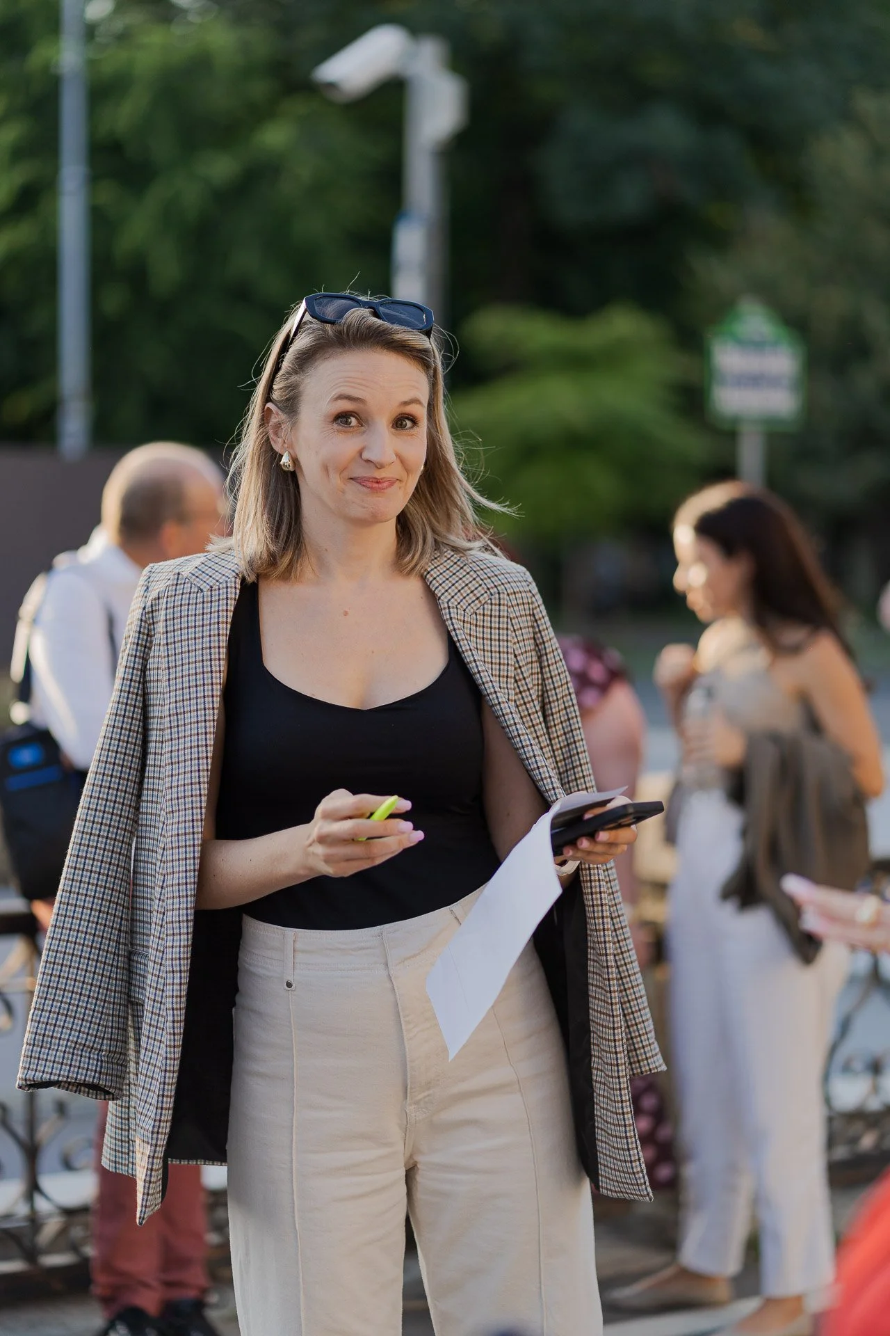 A woman with shoulder-length blonde hair wearing glasses on her head, a checkered blazer, and light-colored pants, holding a phone and some papers, standing outdoors with people in the background.