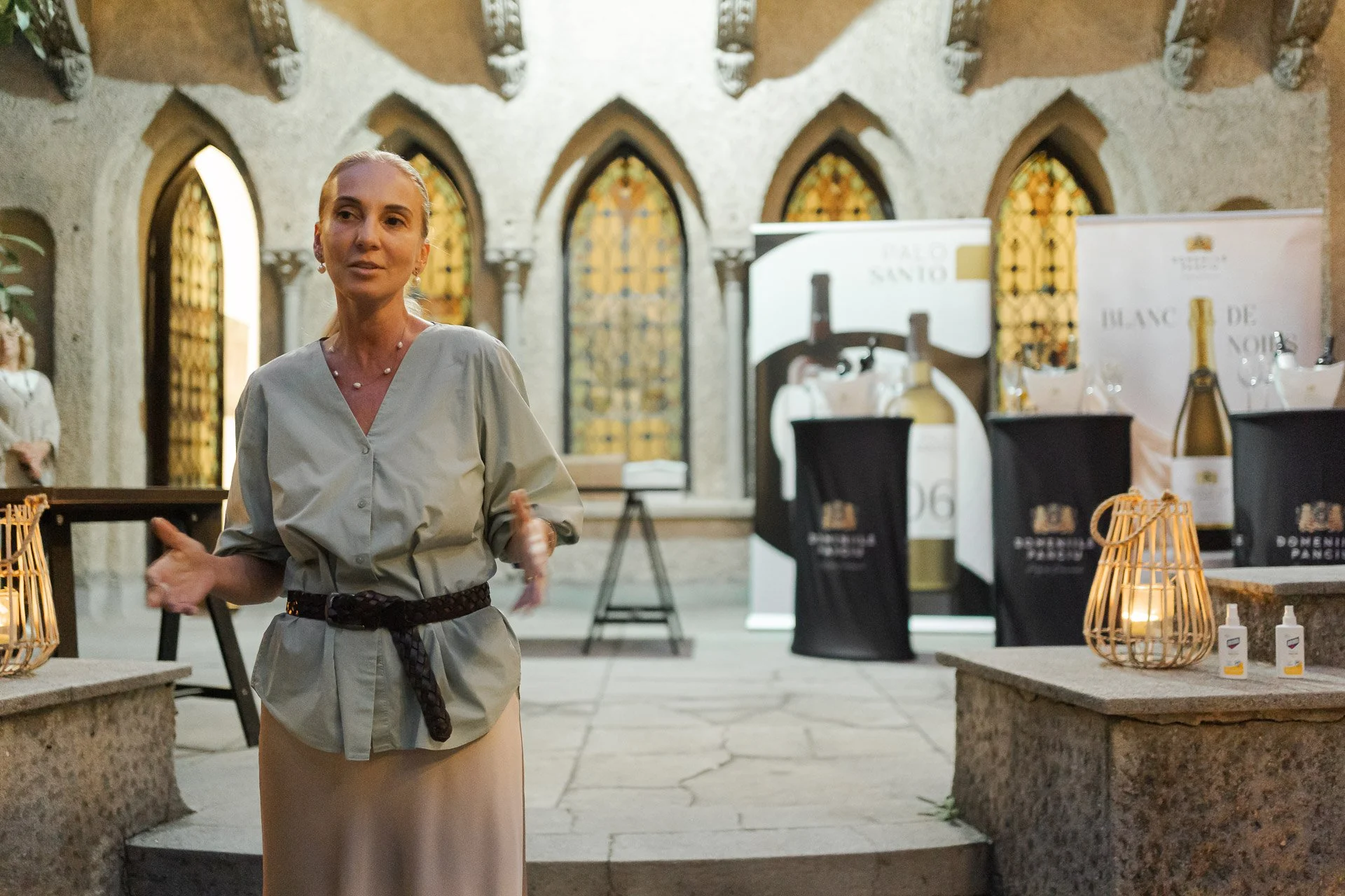 A woman in a beige dress and a black belt stands inside a building with arched stained glass windows, speaking to a group. There are banners with bottles and wine-related graphics in the background, and a table with candles and sanitizer on either si