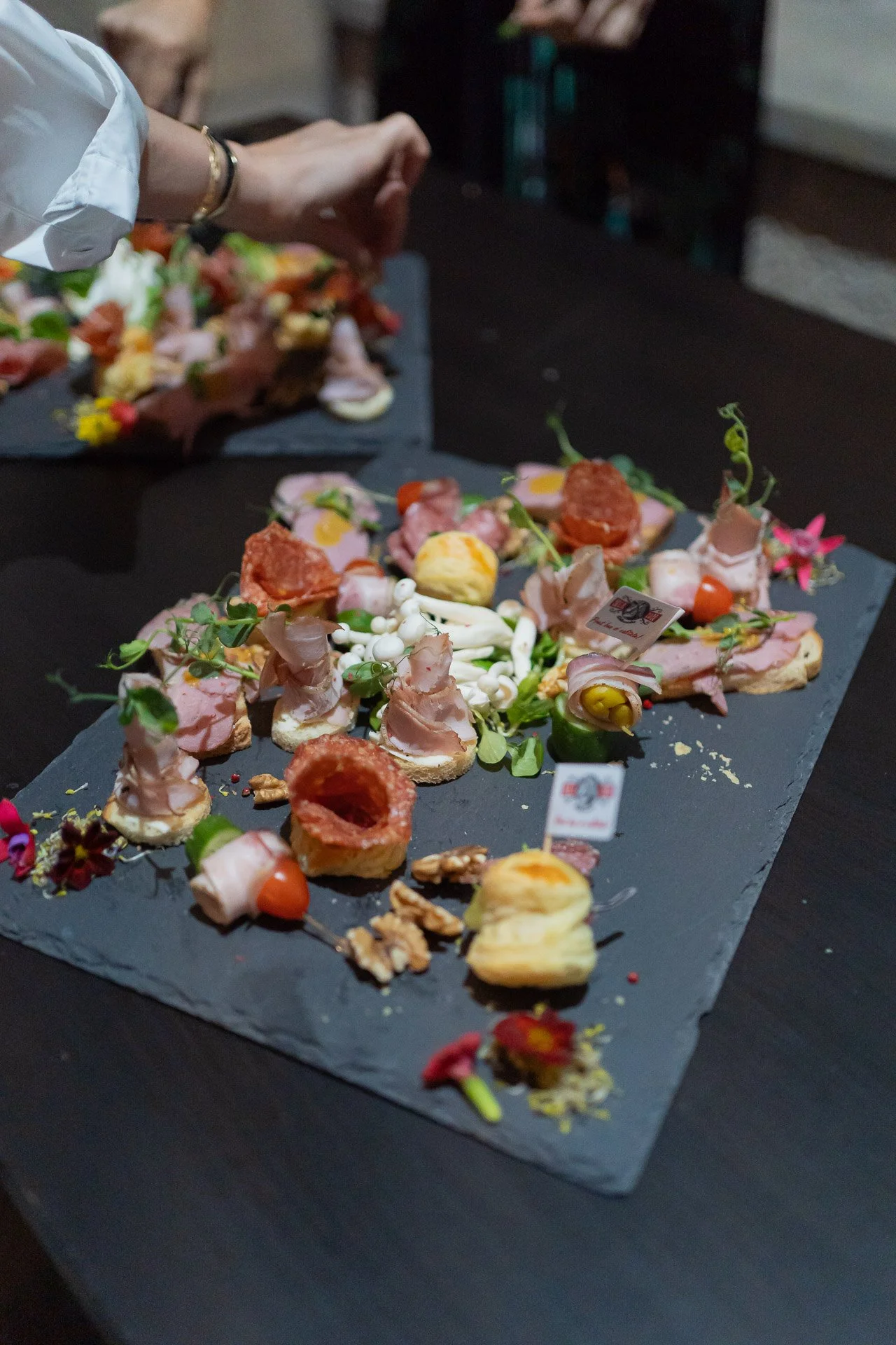 Assorted gourmet appetizers on a black slate platter, including slices of ham, cherry tomatoes, cheese, nuts, and garnishes, with a person arranging them in the background.