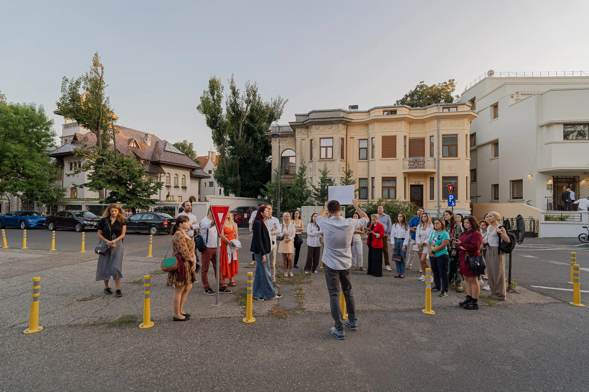 Group of people gathered outdoors during late afternoon or early evening, some listening to a speaker holding a blank sign, surrounded by parked cars and elegant residential buildings.