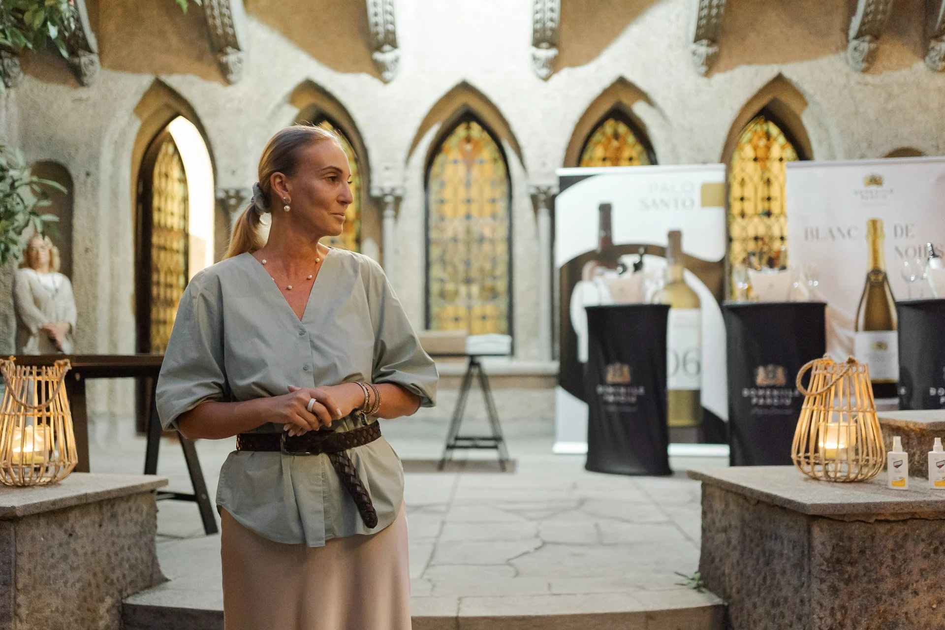 A woman in a gray top and beige skirt, with jewelry and a belt, standing in an ornate church or cathedral with arched windows and banners in the background.